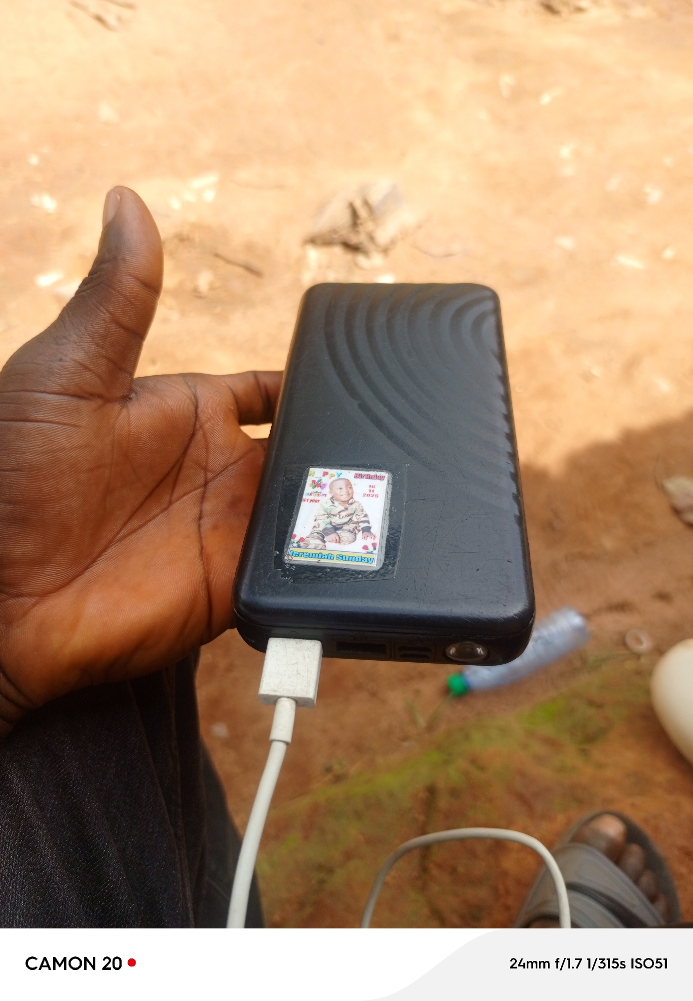 This media file shows a close-up of a person's hand holding a black power bank. A small sticker on the power bank displays a picture of a baby and the text "HAPPY Birthday 1 year 16 11 2025 Jeremiah Sunday." A white USB cable is plugged into the power bank, with the other end disappearing out of frame. The background is a blurred image of dry, sandy ground with some patches of green vegetation. A partially visible plastic water bottle with a green cap and a white cap is also in the background. The image appears to be taken outdoors in daylight, with the sandy terrain suggesting a dry, possibly rural or unpaved area. The overall mood is casual and everyday, focused on the power bank and its sticker. The text at the bottom of the image reads "CAMON 20 24mm f/1.7 1/315s ISO51", indicating the device used to capture the photo and its settings.