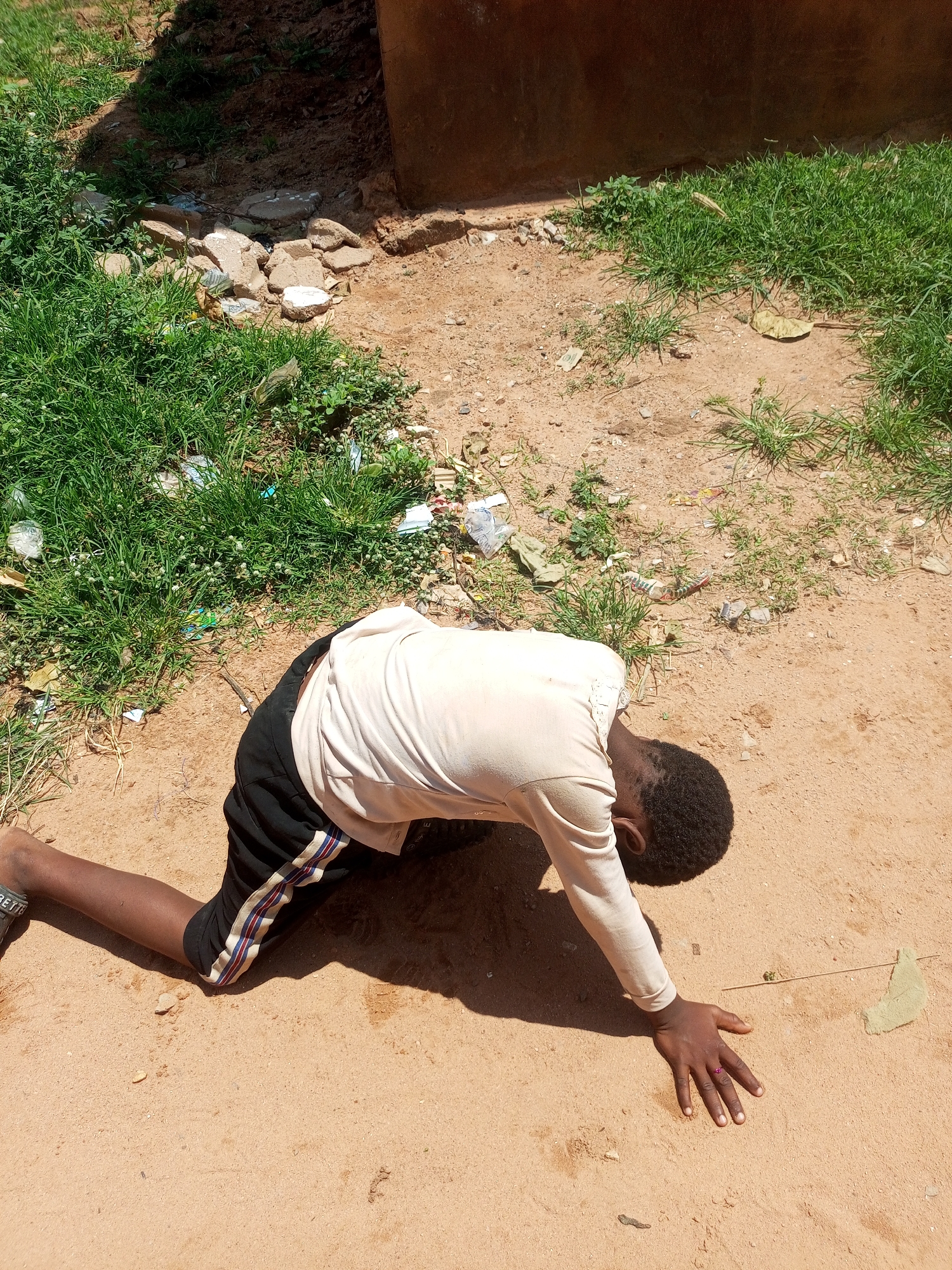 A young Black male is kneeling on sandy ground in what appears to be an outdoor urban environment in Tempure, Nigeria. He is facing downwards, with his head bowed towards the ground. He is wearing a light-colored long-sleeved shirt and black shorts with red and white stripes on the sides. His right hand is extended on the ground, and he is wearing a small pink ring on his right ring finger. The surrounding area is a mixture of dirt, sparse green grass, and some scattered debris, including plastic wrappers and small stones. A brown wall is visible in the background. The scene suggests a moment of prayer, reflection, or perhaps distress.