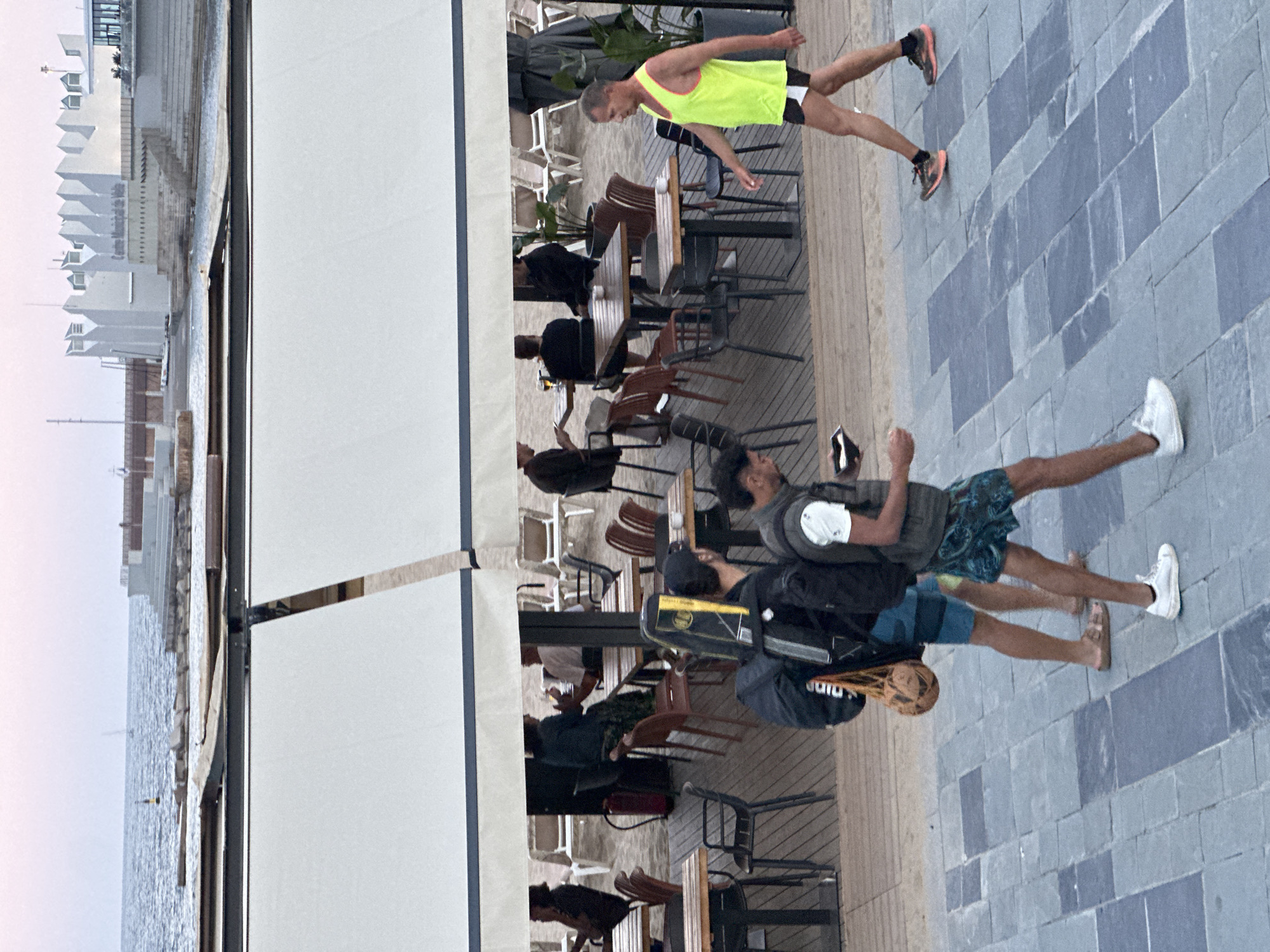 The image depicts a seaside promenade and outdoor dining area, likely in Barcelona, Spain, given the architectural style of the building in the background which bears the inscription "Centre Municipal de Vela". The scene is set against a backdrop of the Mediterranean Sea and a modern building complex.
In the foreground, two individuals are walking along a paved walkway with a patterned tile design. The person on the left carries a large backpack and what appears to be a surfboard case over their shoulder. They are wearing blue patterned swim shorts and white sneakers. The person on the right is wearing a black vest over a white t-shirt and patterned swim shorts.
Further back, under a white awning that covers an outdoor seating area, several people are seated at tables on a wooden deck. Some are dining or conversing. Beach chairs are visible in the background on the sand. A man in a bright yellow tank top and black shorts is walking on the promenade in the mid-ground, towards the right side of the frame. He is wearing running shoes.
The environment suggests a public recreational area, possibly a beach club or restaurant adjacent to a marina. The lighting indicates it is either early morning or late afternoon, with a soft, diffused light.