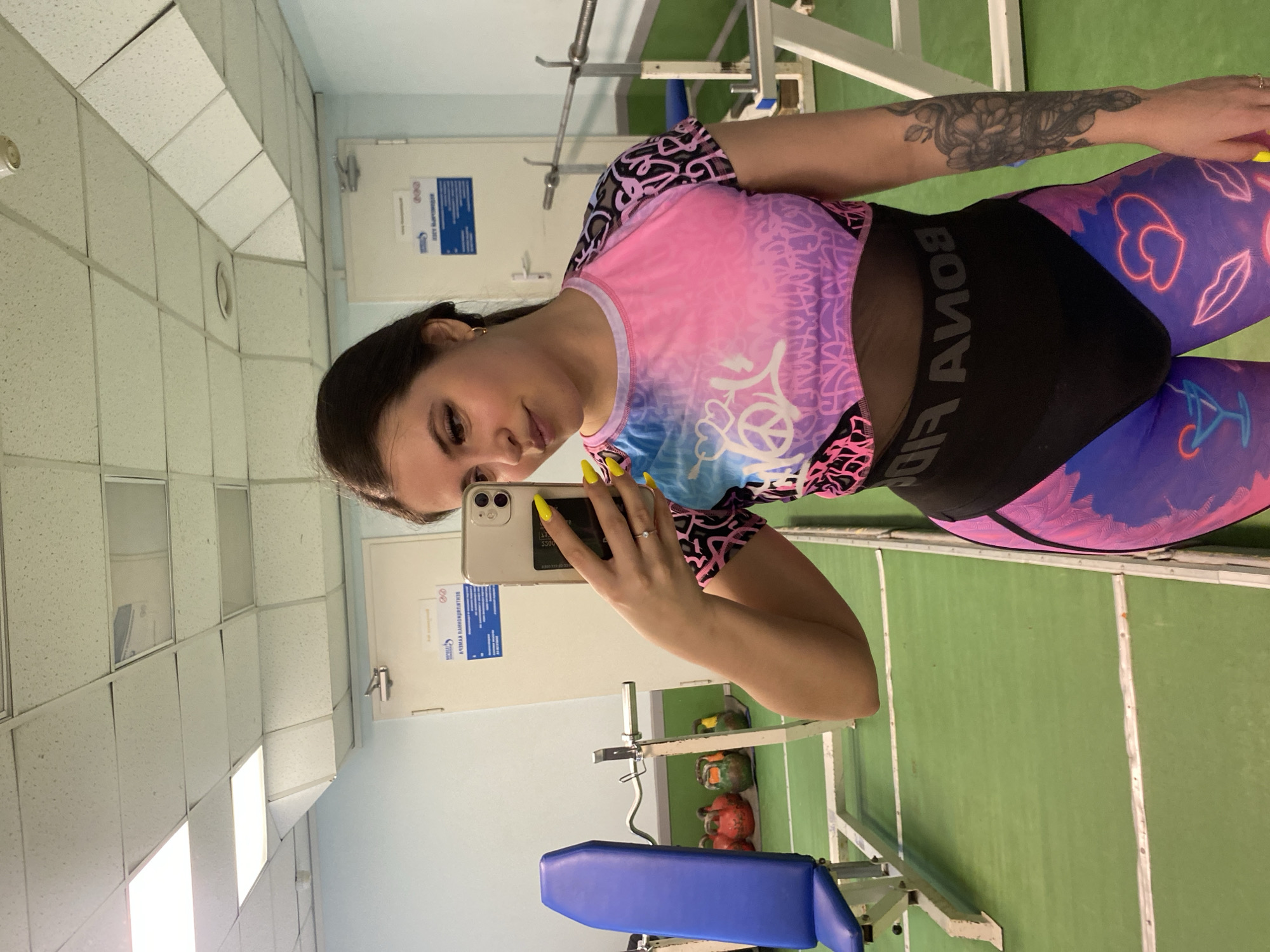 A woman is taking a selfie in what appears to be a gym. She is wearing a pink and blue patterned top with black accents, as well as patterned leggings. Her left arm is visible and has a tattoo. She is holding a smartphone. The background shows gym equipment and a light blue wall. A sign with text in Russian is on the wall, suggesting a location in Russia, possibly Chaikovsky. The ceiling is tiled. The floor is green with white lines.