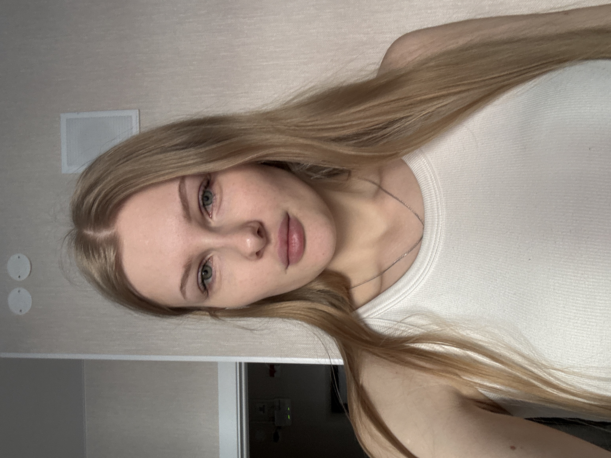 The image presents a close-up portrait of a young woman with long, straight, golden blonde hair that falls over her left shoulder and chest. She has fair skin, light-colored eyes (appearing greenish-grey), and full, naturally pink lips. Her expression is neutral to slightly pensive as she looks directly at the camera. She is wearing a light-colored, possibly white or off-white, ribbed tank top and a thin silver-toned chain necklace.
The background indicates an indoor setting with plain, light-colored walls, likely beige or off-white. On the wall behind her head, a small rectangular white object, possibly an air vent or speaker cover, is visible. Further to the right, two circular white covers, potentially for electrical outlets or similar fixtures, are mounted on the wall. Below her right shoulder, a dark area suggests another part of the room or a reflection, with faint green lights indicating the presence of electronic devices. The lighting is soft and even, characteristic of indoor artificial light. There is no discernible text within the image. The scene suggests a posed photograph, possibly a selfie, taken indoors in Perm, Russia.