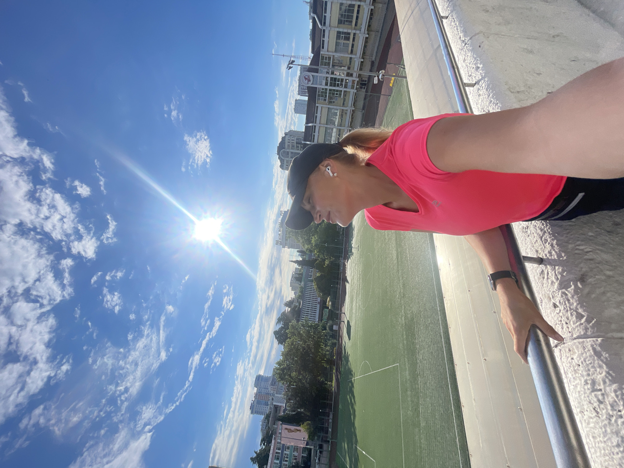 A woman is taking a selfie on a sunny day with a bright blue sky and some clouds, looking up towards the sun. She is wearing a pink t-shirt, a black baseball cap, and white wireless earbuds. Her right hand is on a metal railing in the foreground. In the background, there is a green sports field with white markings, likely a soccer or football field, surrounded by buildings and trees. The overall scene suggests an outdoor sports facility in an urban setting. The location is identified as Sochi, Russia.