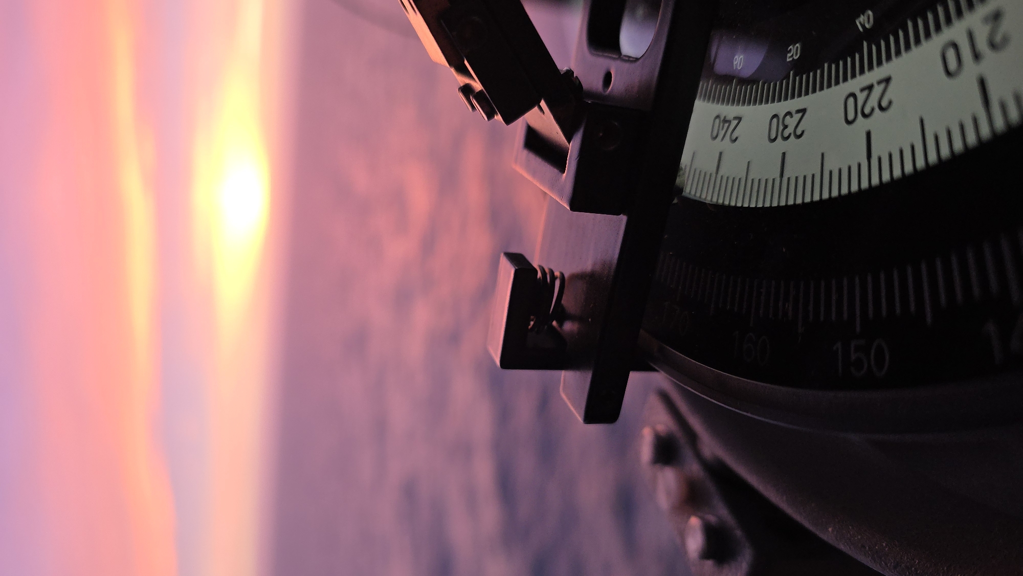 The image shows a close-up of a navigation instrument, likely a sextant, with a view of a sunset in the background. The instrument's scales are visible, showing markings from 140 to 240. The sunset features a bright yellow sun and a pink and purple sky. The image does not provide enough information to determine the location or city.