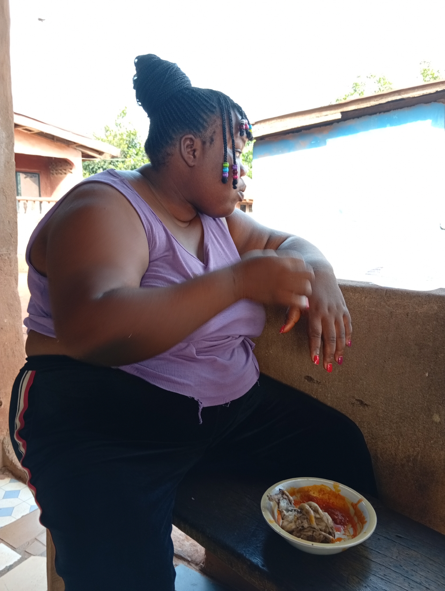 A medium shot depicts a woman seated on a wooden bench in Ogidi, Nigeria. She wears a lavender tank top, dark pants with a red and white stripe, and has her hair braided and styled in a bun with colorful beads. Her nails are painted red and pink. She rests her left arm on a tan wall or ledge. A white bowl containing food is placed on the bench near her. The background includes a glimpse of buildings with blue and brown exteriors, and green foliage is visible above. The image is well-lit, and appears to be taken during the daytime.