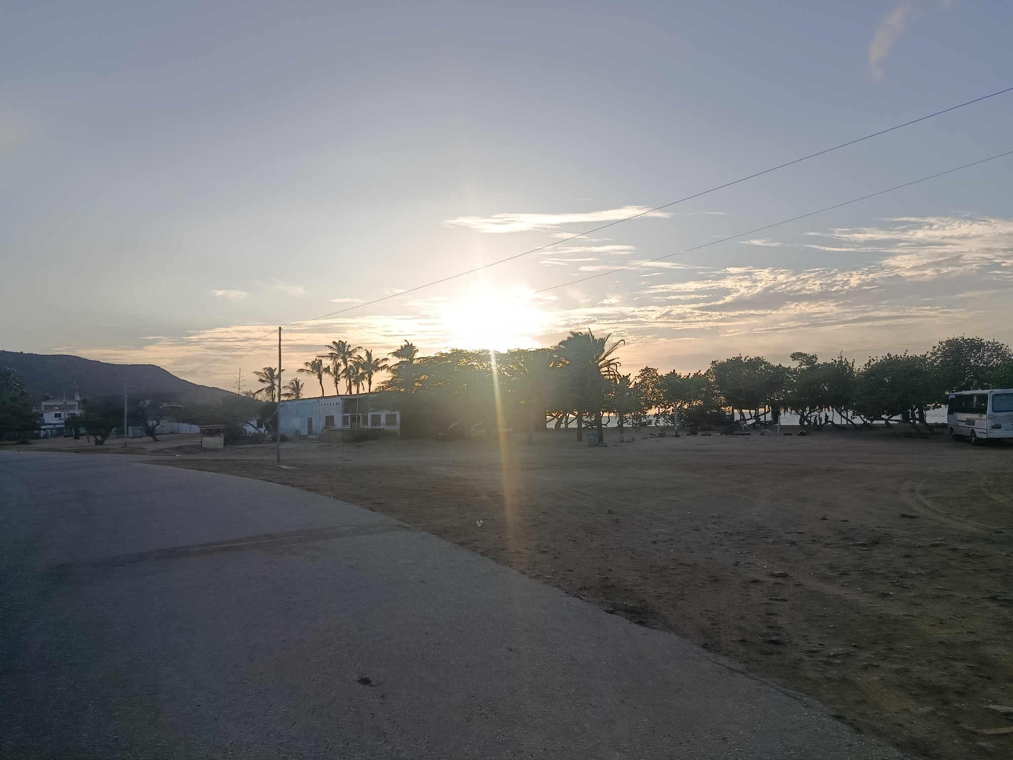 The image depicts a view in Altagracia, Venezuela at sunset. The foreground contains a paved road on the left, transitioning to a sandy area on the right. Trees, including palm trees, are silhouetted against the setting sun, which casts a strong glare. A building is visible behind the trees, as is a bus parked on the right side of the image. In the background, a hill or small mountain is seen on the left. Overhead, power lines stretch across the sky. The sky is a mix of blue and white, with scattered clouds.