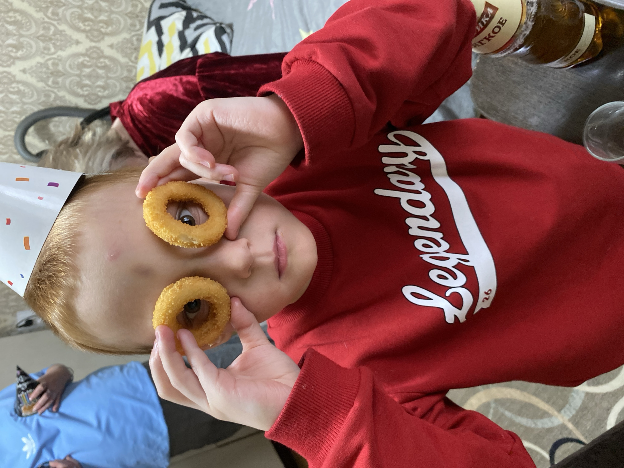 A young boy, wearing a red "Legendary" sweatshirt and a birthday hat, holds two onion rings in front of his eyes. He is indoors. To the right of him is a bottle of alcohol. In the background, a person wearing a blue shirt is visible. Based on the label on the bottle, the location is Chaikovsky, Russia.