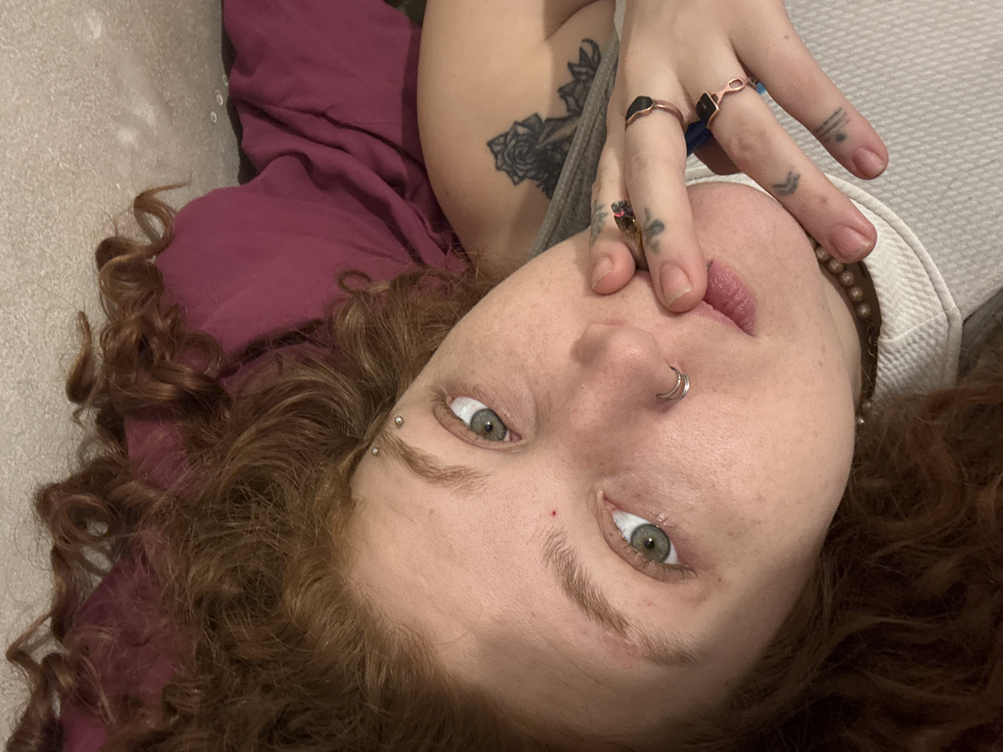 An intimate, close-up, and inverted portrait, likely a selfie, features a person lying down and looking directly into the camera. The individual, situated in Buenos Aires, Argentina, has fair skin, striking greenish-gray eyes, and a cascade of curly, reddish-brown hair framing their face. Their expression is direct and engaging, with a hint of a soft smile as their fingers lightly touch their lips and nose.
Notable details include multiple facial and body adornments. A silver septum ring is visible, along with two horizontal eyebrow piercings. Several rings with dark stones are worn on their fingers, which also bear small, intricate tattoos, including line patterns, a chevron, and a cross-like symbol. A larger floral tattoo, possibly a rose, is visible on their upper arm. A beaded necklace is partially seen around their neck.
The person is resting against a maroon-colored fabric, likely a blanket or clothing, which contrasts with a lighter, textured surface in the background. This arrangement suggests an indoor setting, such as a bedroom or living room. The lighting appears soft and ambient, without strong shadows, indicating an indoor shot, possibly during the day or evening. No visible text is present in the image. The overall mood is relaxed and personal, conveying a direct connection with the viewer.
