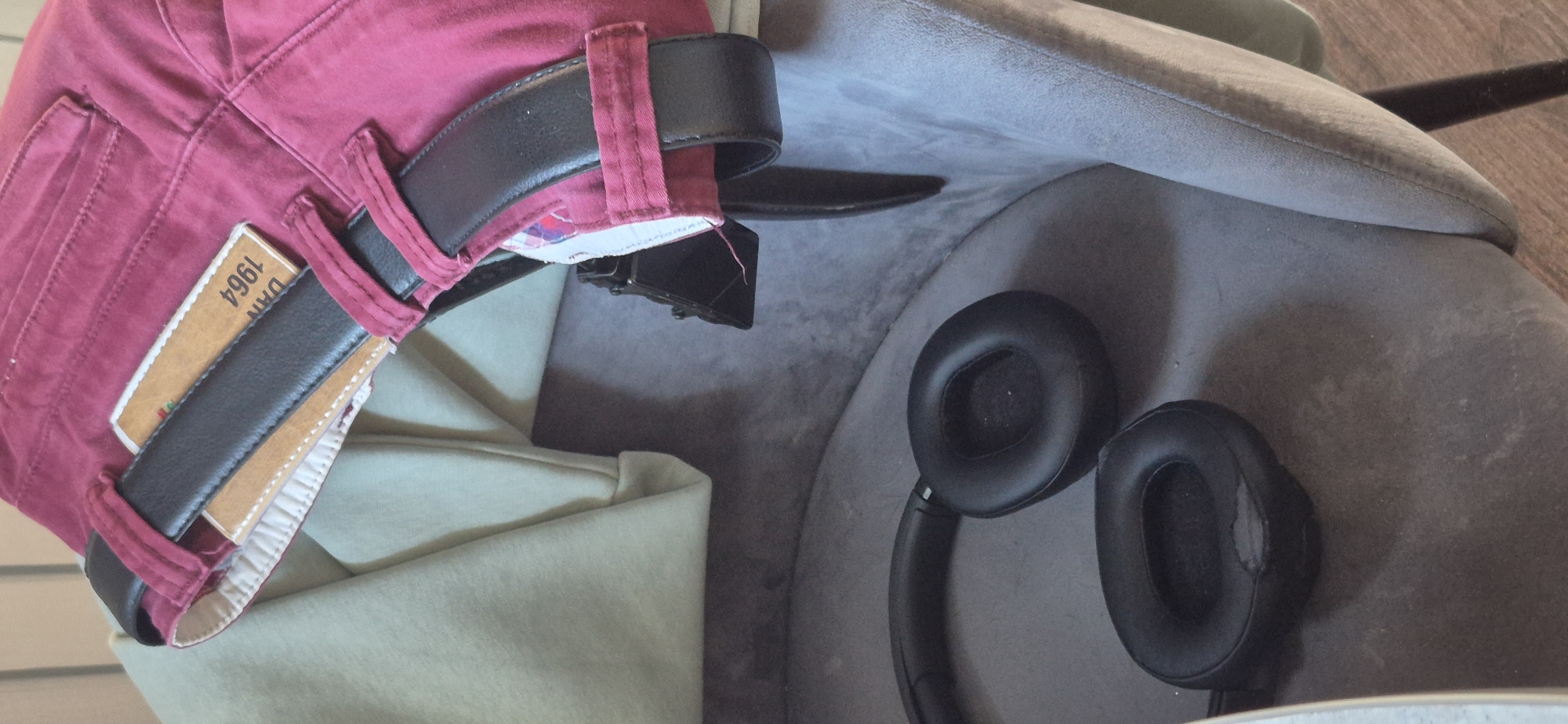 The image shows a pair of black headphones inside a gray chair. Red trousers with a brown leather belt are draped over the chair. The label on the trousers reads "DAN 1964". The chair is placed on a wooden floor. The city is unknown.