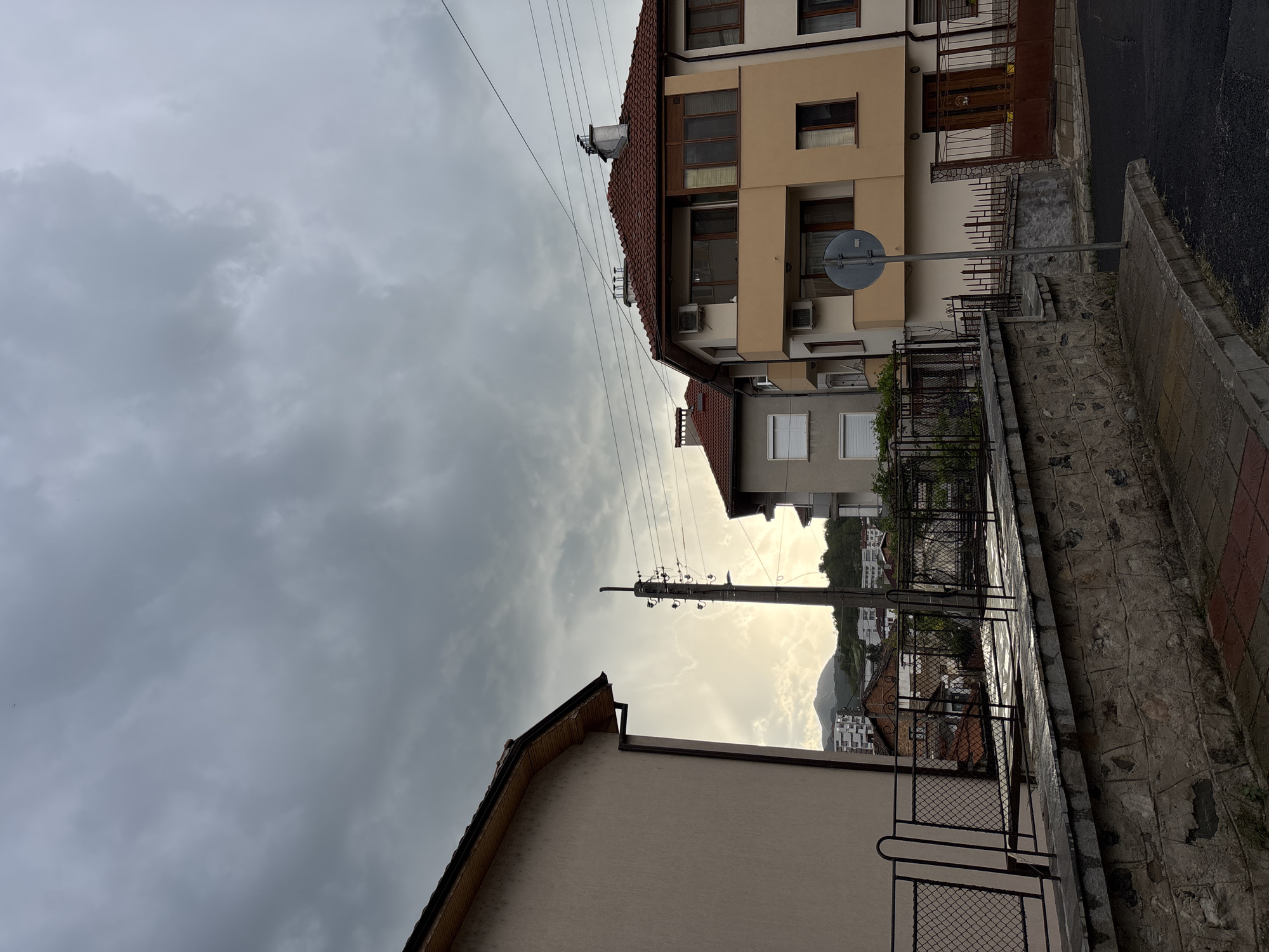 The image depicts a cloudy sky over the city of Zlatograd, Bulgaria, with a view down a narrow street. On the right is a multi-story building with a red tile roof, beige facade, and several windows. A road sign is attached to the building. A stone retaining wall runs along the street. On the left is a building with a tan-colored facade. Power lines are visible overhead. In the distance, a mountain is visible in the bright sunlight, partially obscured by buildings.