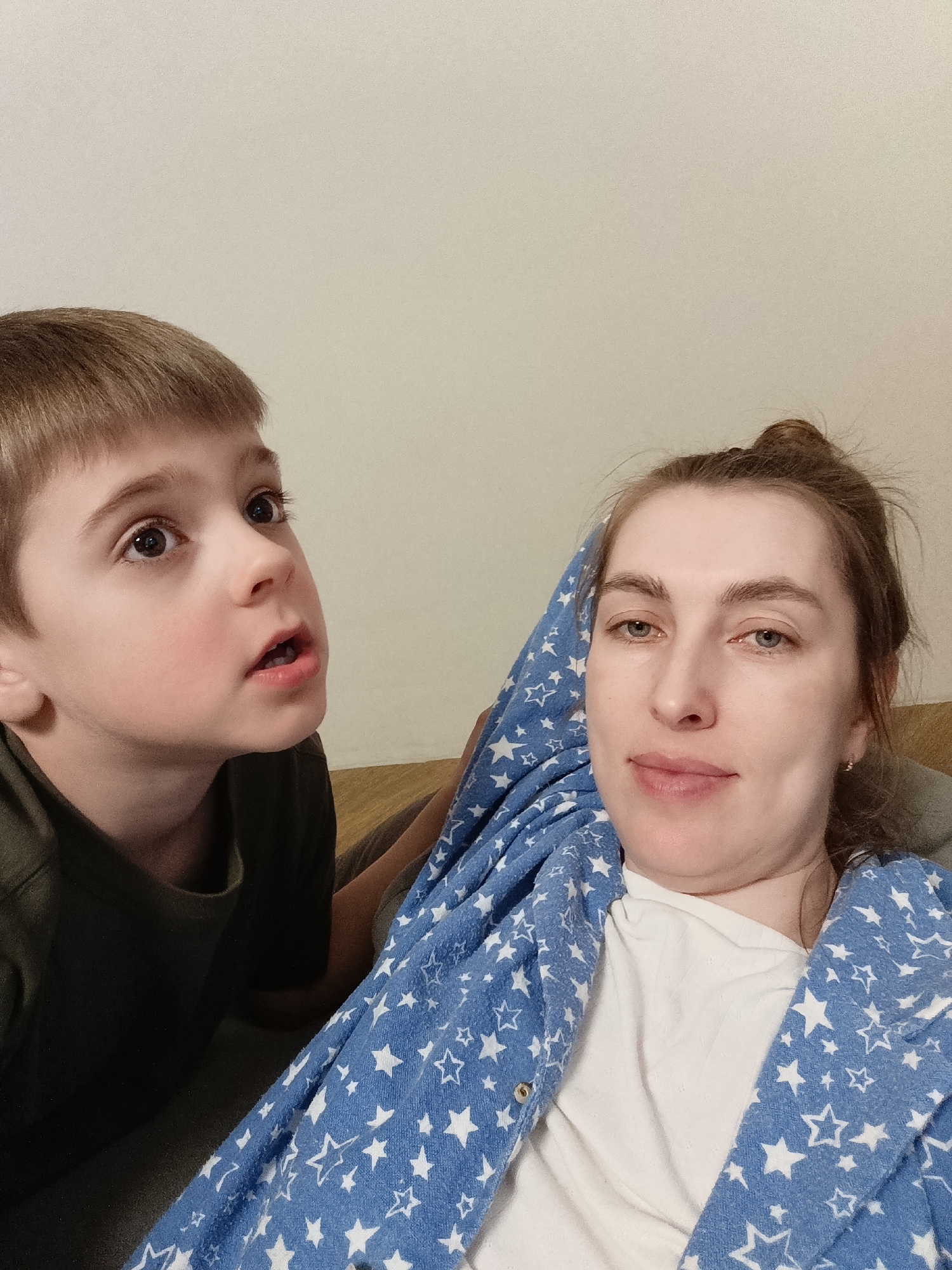 A woman and a young boy are in what appears to be an indoor setting. The woman is wearing a white shirt and a blue robe with white stars. Her hair is up in a bun. The boy is next to her and wearing a dark shirt. The background appears to be a plain, light-colored wall. No indication of the location is present.