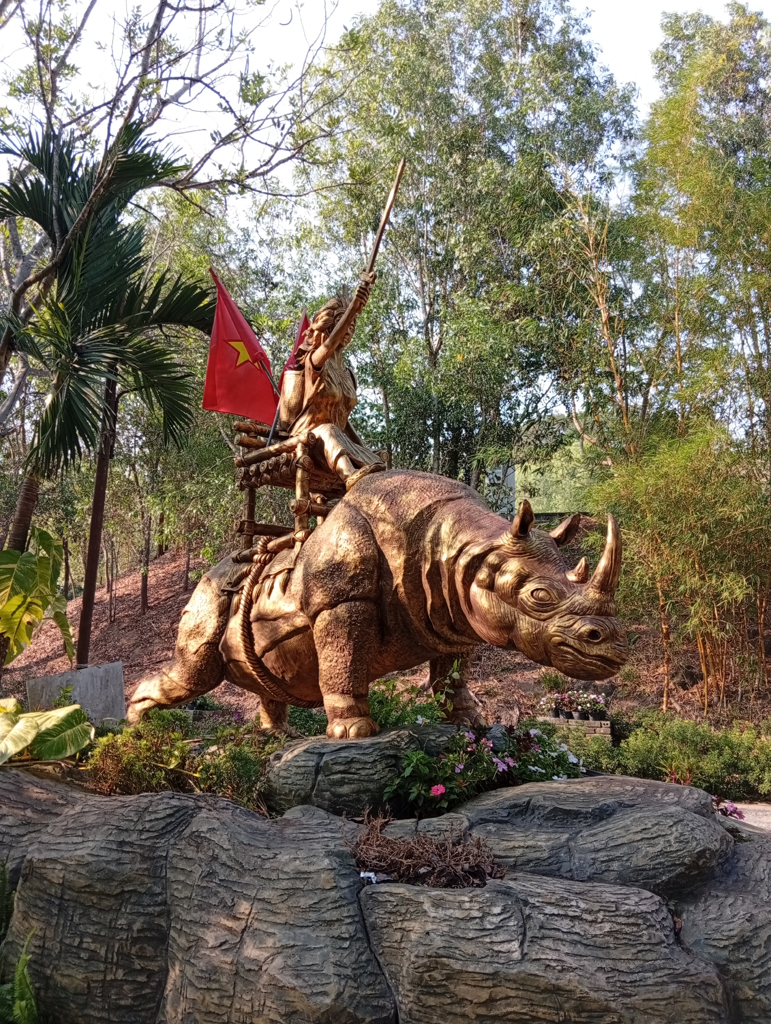 The image displays a prominent golden statue of a warrior figure riding a rhinoceros, set against a backdrop of lush greenery. The warrior, possibly female, is depicted with long, flowing hair and traditional attire, holding a long spear or staff aloft in a victorious pose. Behind the warrior, a vibrant red flag with a yellow star, the national flag of Vietnam, is affixed to the saddle structure of the rhinoceros.
The rhinoceros statue is rendered in a detailed, golden-bronze hue, showcasing its powerful build, textured skin, and prominent horns. Both figures convey a sense of dynamic movement and strength. The statue is mounted on a base of large, textured grey artificial rocks, around which various small flowering plants (pink, white, purple) and green foliage are arranged.
The background consists of dense trees and tropical vegetation, including palm trees on the left and bamboo on the right, suggesting a forest or heavily landscaped garden setting. A bright sky is visible through the tree canopy. The lighting indicates it is daytime, with clear weather, as sunlight illuminates the golden statue and surrounding plants. The overall scene evokes heroism, national pride, and historical or mythological significance. The location context provided is Tân Phú, Vietnam. No visible text is present in the image.