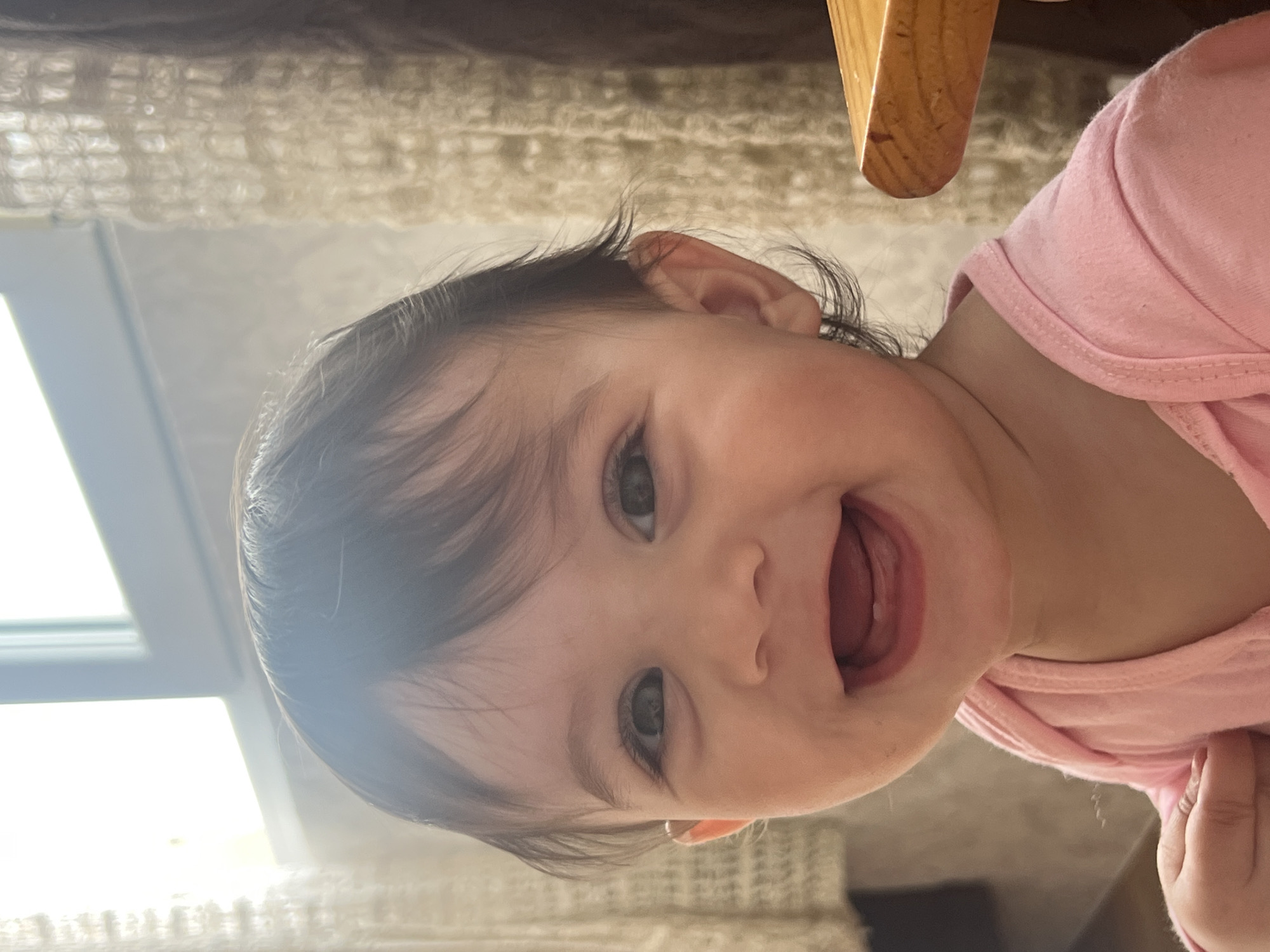 A close-up portrait of a baby with dark hair and light eyes, wearing a pink shirt, is captured in a brightly lit environment. The baby's mouth is open, possibly smiling or speaking. A wooden object is partially visible in the upper right corner of the frame. The location is Elena, Bulgaria. A window with light filtering through is seen on the left side of the image.