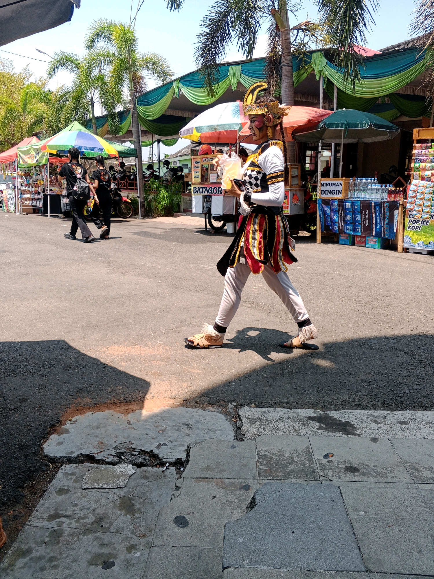 In the city of Wuryorejo, Indonesia, on a paved road under a clear, sunny sky, a person dressed in traditional attire walks from left to right. The individual wears a gold headpiece with intricate designs, makeup on their face, a white long-sleeved top with black and white checkered details on the chest, and a layered skirt of red, yellow, and black fabric. They also have light-colored pants and tan sandals. The person is holding a clear plastic cup filled with a yellow liquid. Behind the individual, there are several market stalls with colorful awnings. One stall displays drinks, including bottled water and other beverages, with signage that reads "MINUMAN DINGIN" (Cold Drinks). Another stall has various snacks or merchandise. Palm trees and other foliage are visible in the background. The ground in the foreground is asphalt, with a section of cracked and broken concrete pavement. Two other individuals, dressed in black, are walking away from the market stalls in the background. Motorbikes are parked near the stalls.