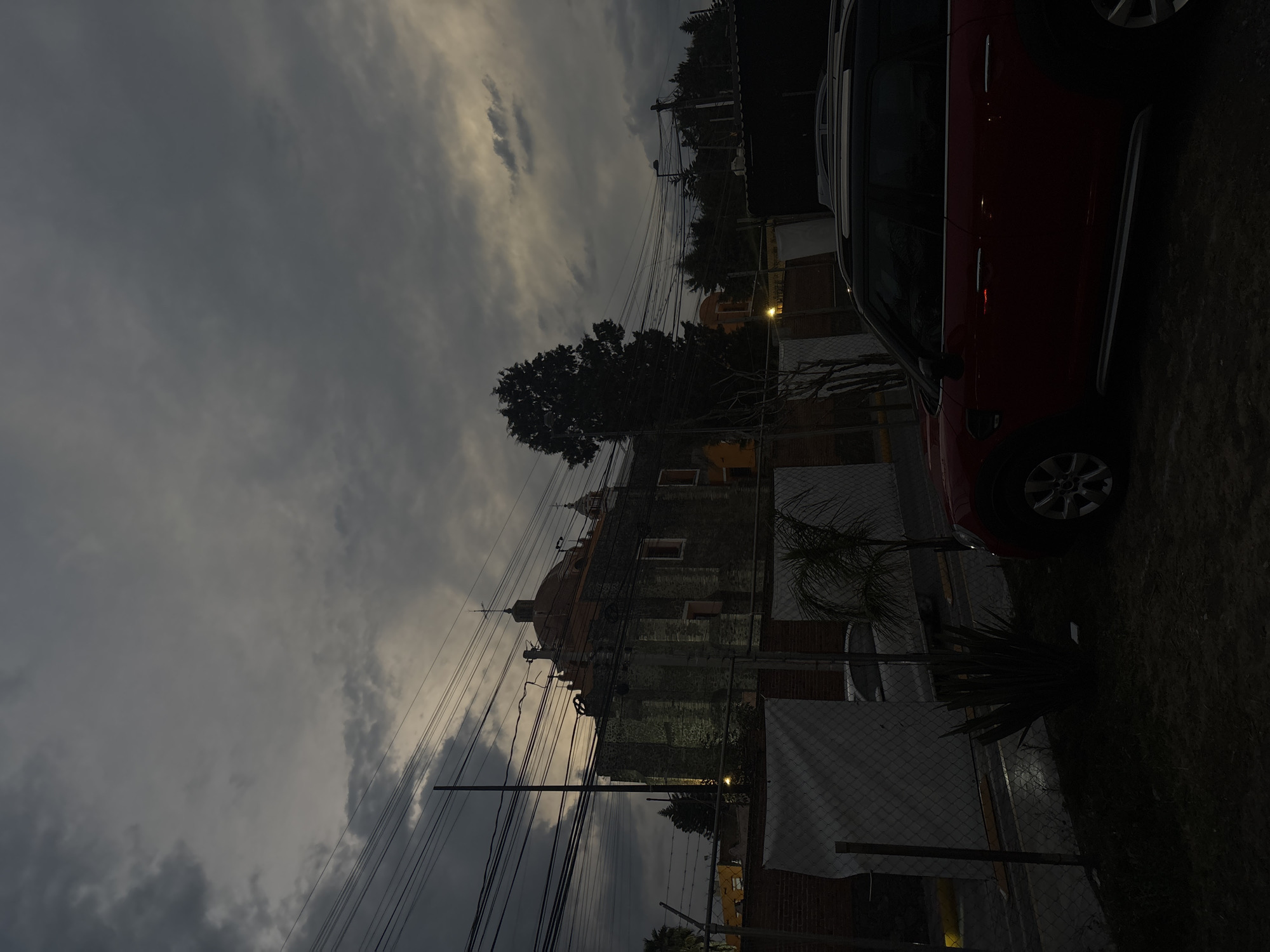 The image depicts a partially obscured view of a building, possibly a church or historical structure, taken in San Andrés Cholula, Mexico. The building is predominantly stone and brick, featuring a dome-like structure, with power lines visible in the foreground. A red car is situated on the right side of the frame, partially out of focus. The sky is overcast. A tree is prominent near the building. A fence is present near the foreground.