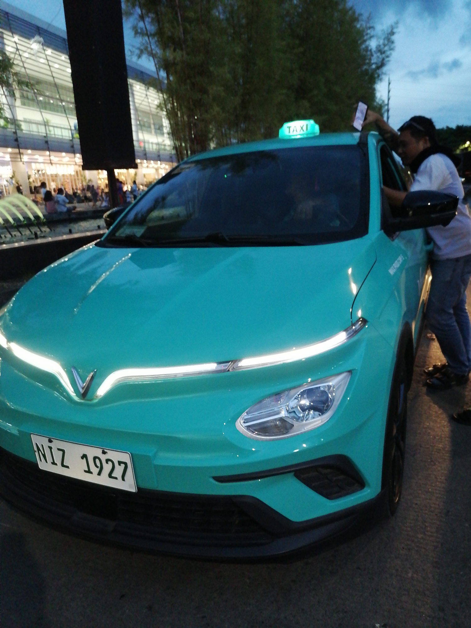 The image depicts a turquoise electric taxi with the license plate "NIZ 1927" parked on a paved surface. A person, wearing a white t-shirt and blue jeans, is standing next to the driver's side door, holding a smartphone. The taxi has a glowing green "TAXI" sign on its roof. In the background, a modern building with glass facades is visible, along with trees and a crowd of people sitting near a fountain. This scene is consistent with the area near SM Lanang Premier in Davao City, Philippines, a popular urban location for shopping, dining, and leisure activities. The lighting suggests it is either early morning or late evening.