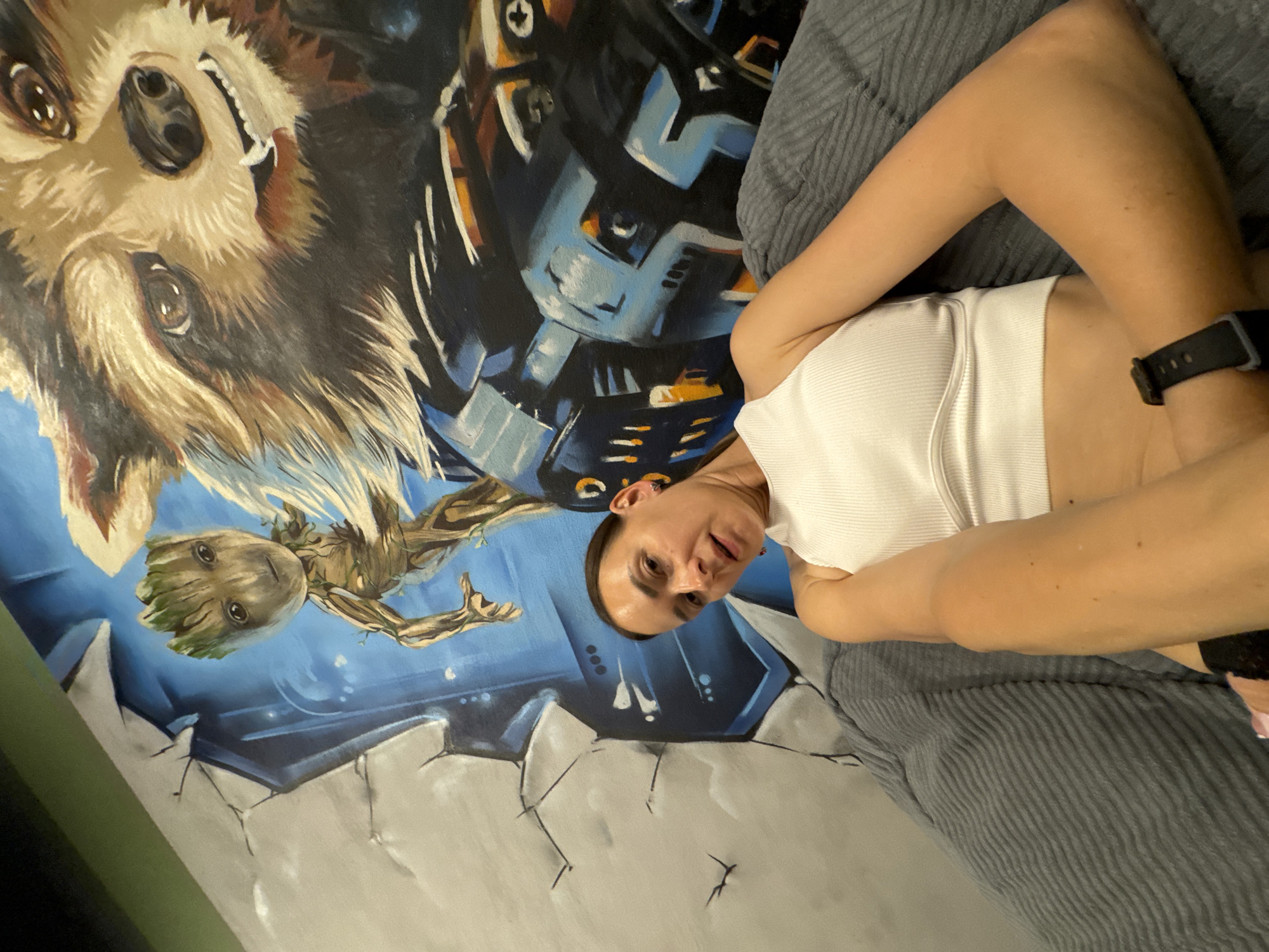 A woman is lying on a gray, ribbed couch in front of a large mural. The mural depicts characters from the movie Guardians of the Galaxy, specifically Rocket Raccoon and Groot. The woman is wearing a white tank top and a black watch. The overall lighting is dim, with some light coming from the left side of the image. There is no specific information about the location in the city of Sochi, Russia.