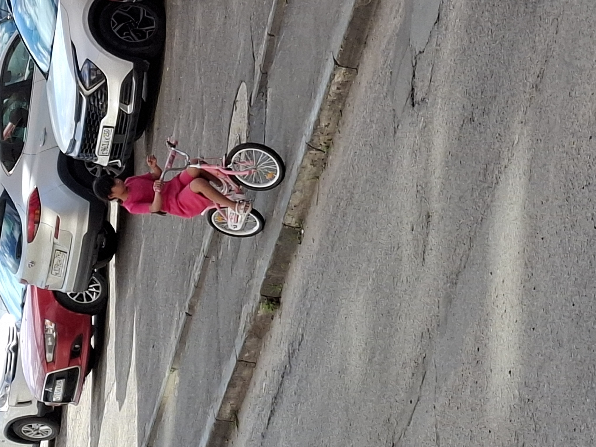 In an urban outdoor setting, a young girl with dark hair, wearing a bright pink dress and sandals, rides a pink bicycle on a paved road. The bicycle has white rims and black tires, with training wheels visible. The girl is positioned in the center of the frame, looking forward. In the background, several cars are parked: a red car in the left foreground, a white SUV behind it, and a white car to the right. The road appears to be on a slight incline with a concrete curb on the left side. The ground is asphalt with some visible cracks and debris. The lighting suggests it is daytime, with shadows present.