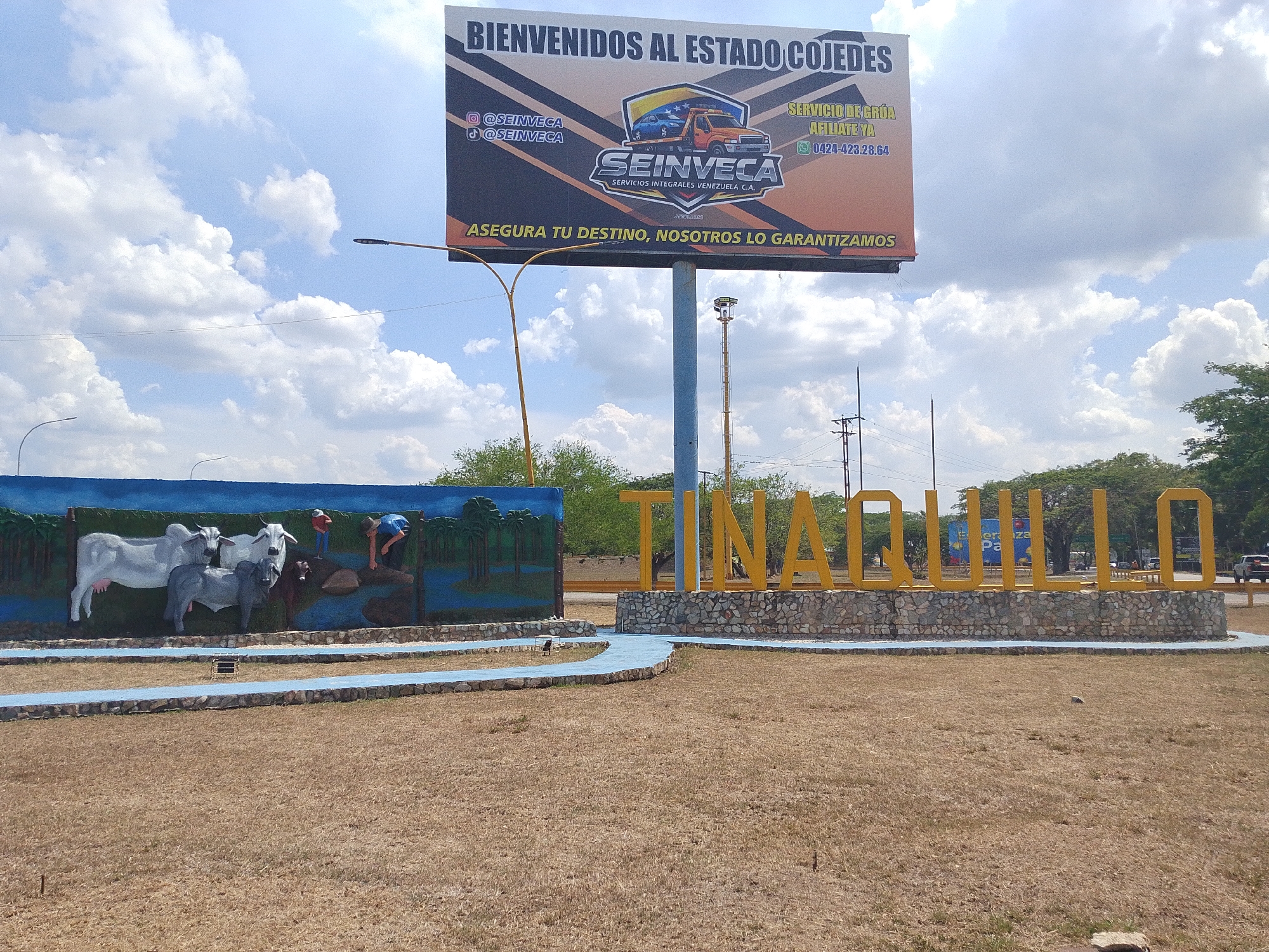 The image is a wide shot depicting the entrance to Tinaquillo, Venezuela. In the foreground is a dry, grassy area. To the left, a sculpted mural depicting cows and people working is visible. To the right, the city's name, "TINAQUILLO," is spelled out in large, yellow, three-dimensional letters, set atop a stone base. In the background, a large billboard for "SEINVECA," a towing service, is seen. The sky is partly cloudy. The image suggests a sunny day in Tinaquillo.