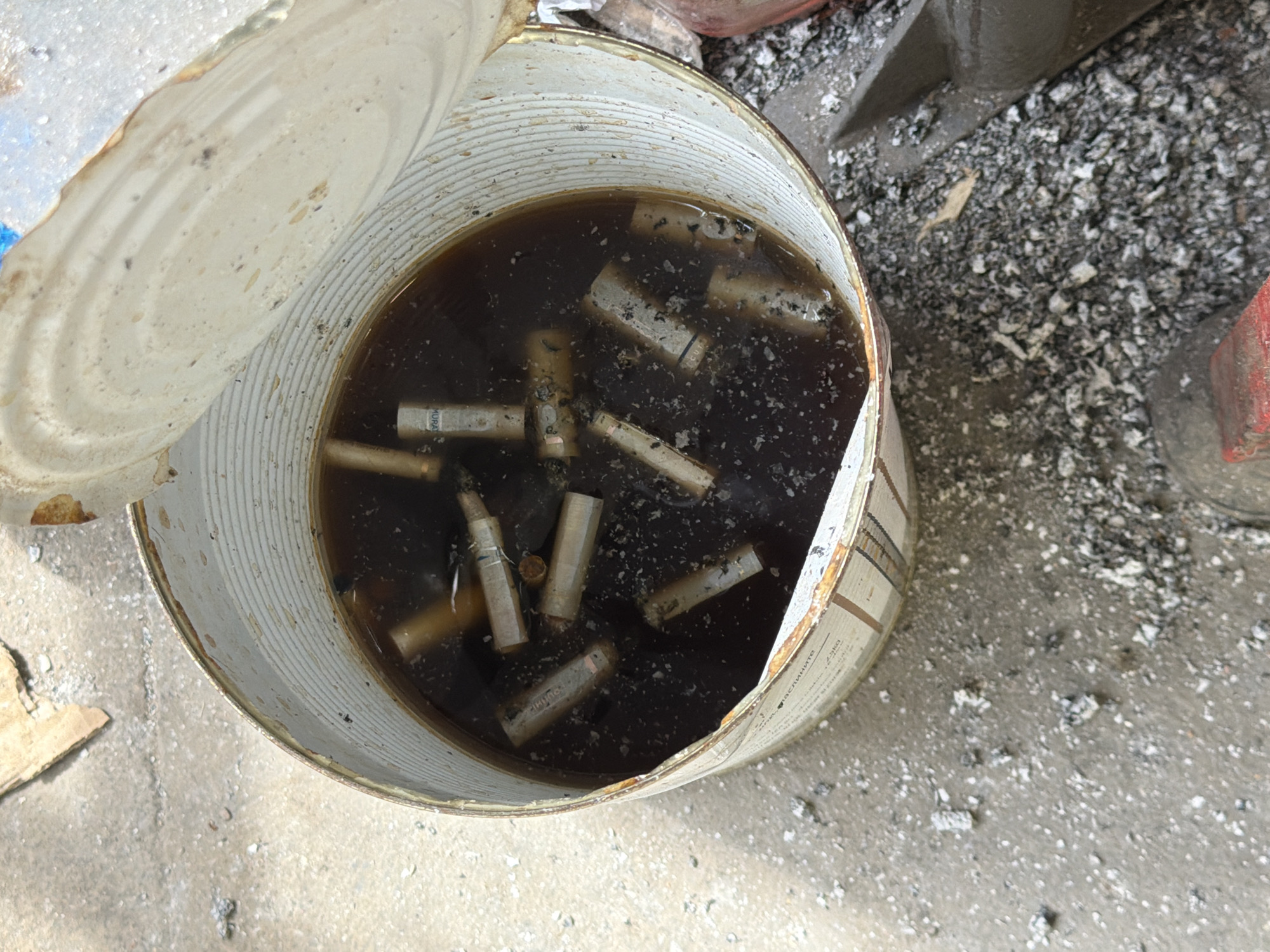 A rusted metal can, with its lid ajar, contains a dark liquid filled with cigarette butts. The can sits on a concrete surface littered with debris, including ash and small stones. To the upper right of the can, there is a pile of crushed material and what appears to be a portion of a brick. The interior of the can is marked by visible rust. The presence of Cyrillic script on the side of the can is visible, suggesting that the scene is in an area that uses the Cyrillic alphabet, specifically the city of Veliko Tarnovo, Bulgaria. No people are visible in the image.