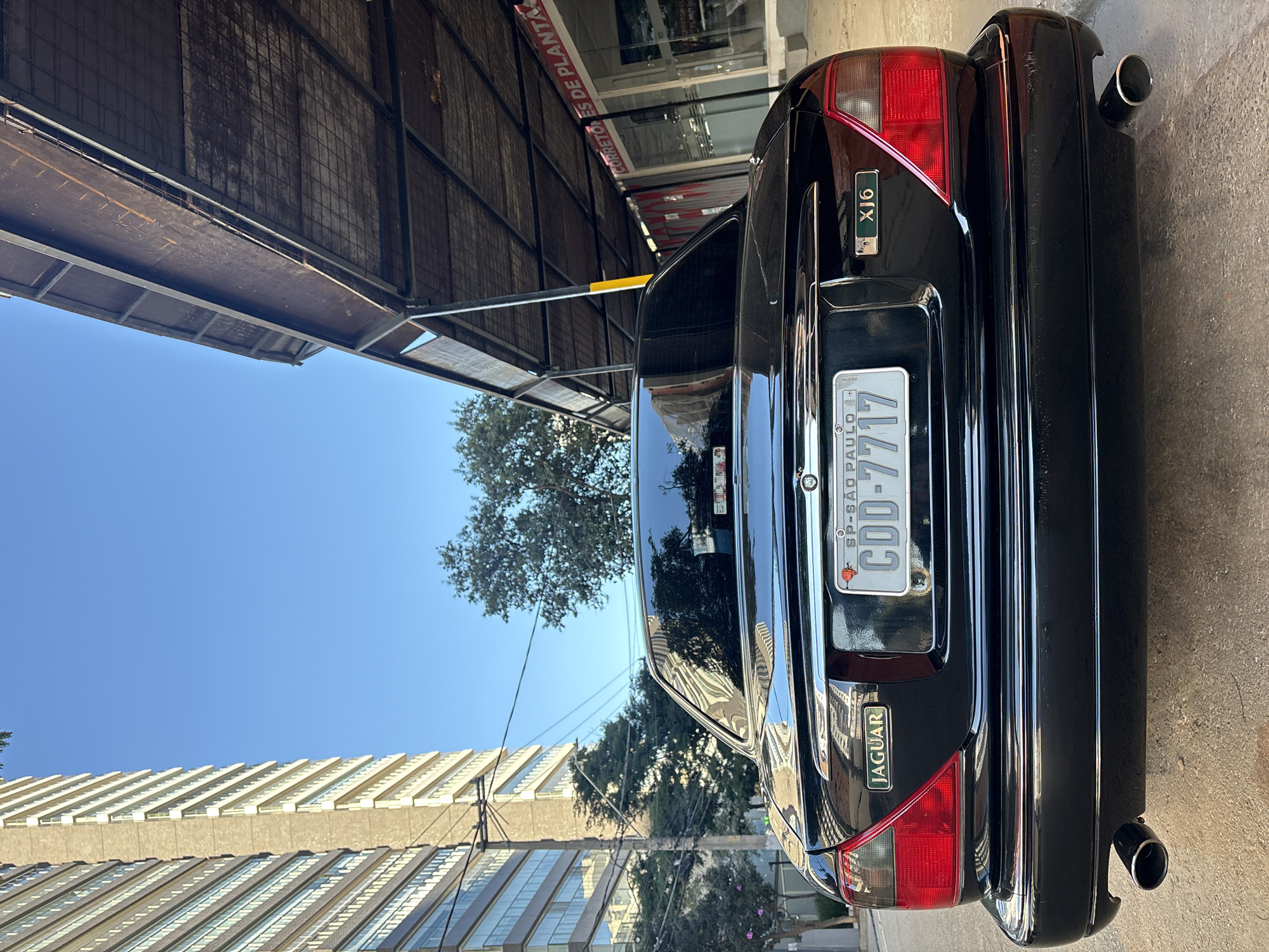 The rear view of a black Jaguar XJ6 sedan is captured in an urban setting. The car is parked on an asphalt surface, with its dual exhaust pipes visible at the bottom. The license plate, reading "CDD-7717" with "SP-SAO PAULO" above it, is centrally located on the trunk. A "JAGUAR" emblem is on the left and an "XJ6" emblem is on the right.
The car is positioned in front of a building with an awning or covered walkway structure. Signs on the building's glass facade are partially visible, with one reading "CORRETORES DE PLANTA". To the left of the car, a tall, modern apartment building with numerous windows and balconies dominates the skyline. Trees are present on either side of the street and behind the car. The sky is a clear, bright blue, indicating daytime. The lighting suggests it's either morning or late afternoon, with the sun casting shadows. The overall scene is typical of a city street in São Paulo, Brazil.