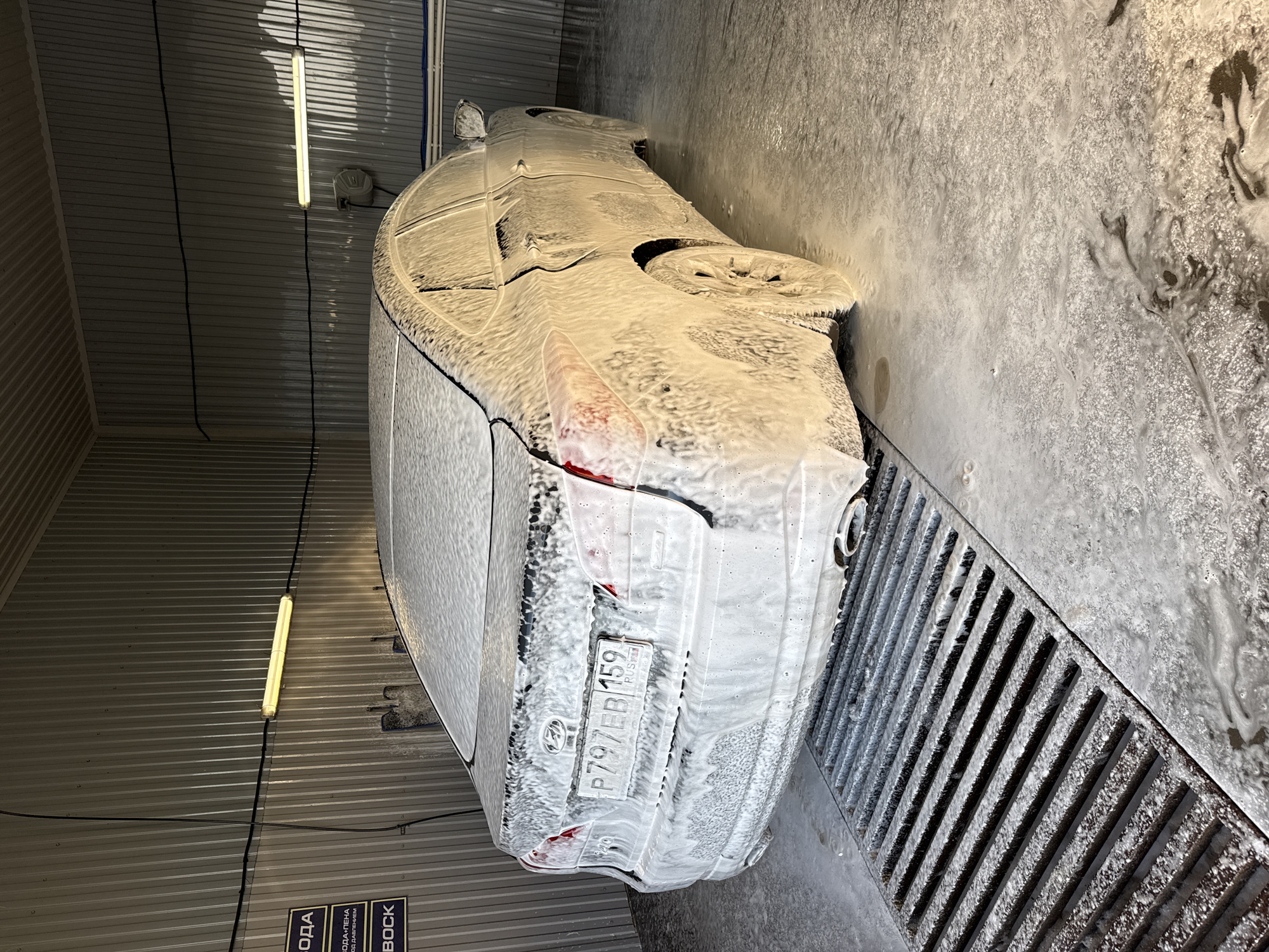 A white car is covered in white foam, seemingly in a car wash facility. The license plate is visible and reads "P797EB 159 RUS." The car is positioned over a metal grate, likely for drainage. The location is suggested to be Chaikovsky, Russia, as indicated by the license plate region code "159." The car wash area appears to be indoors, with metallic walls and fluorescent lighting.