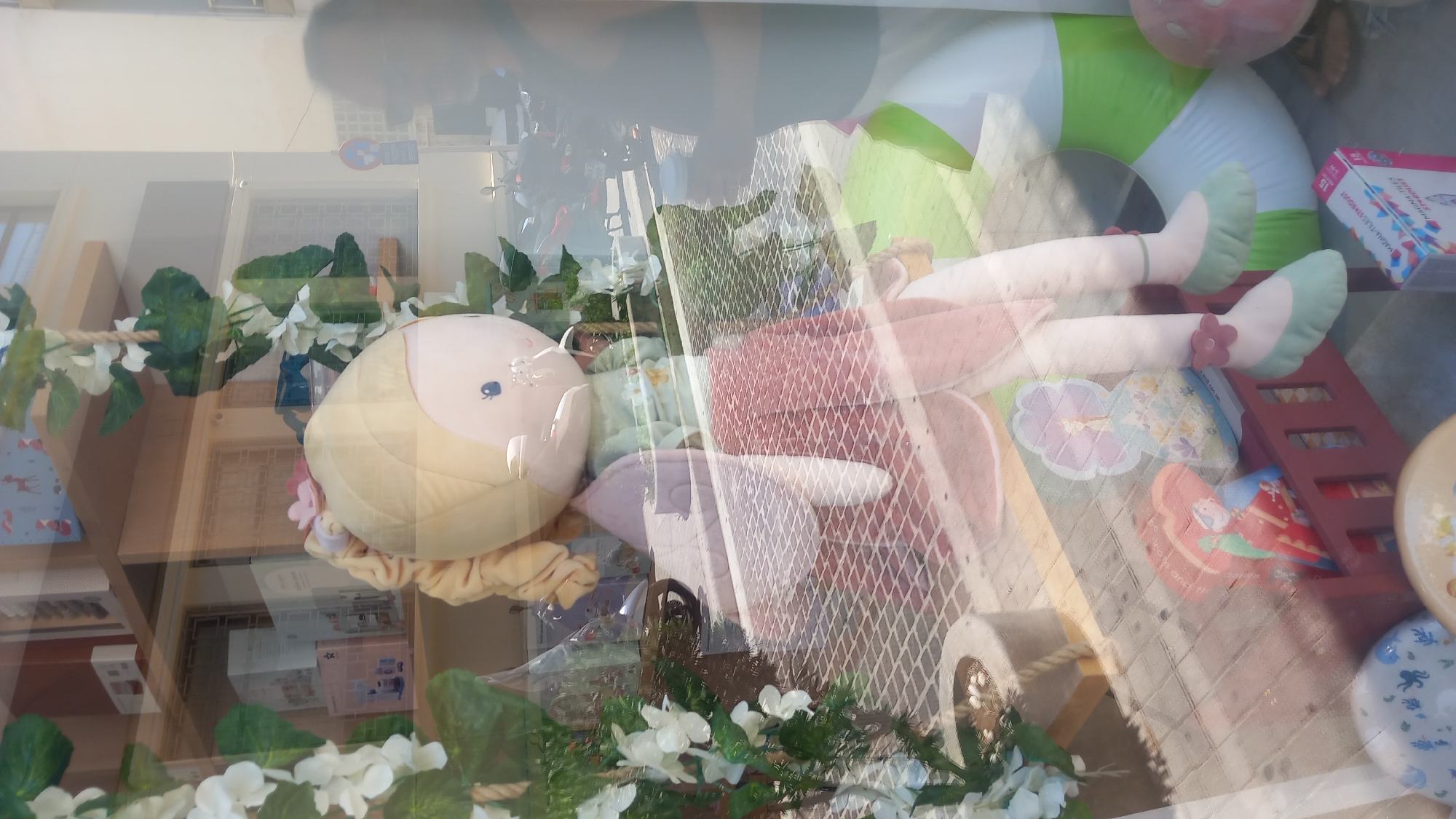 A large plush doll with blonde hair and blue eyes is displayed in a shop window. The doll is wearing a pink dress and light green shoes. It is surrounded by artificial flowers and foliage. In the background, shelves are visible with various items, including boxes and picture frames. Through the window, reflections of a street, buildings, and people can be seen, indicating an urban setting. Specifically, the location is identified as Kos, Greece. There is a partial view of a person in silhouette standing to the right of the doll, also reflected in the window. The flooring inside the shop appears to be a tiled pattern. Several other objects are scattered around the display, including a red wooden toy crib, an inflatable ring, and some decorative boxes.