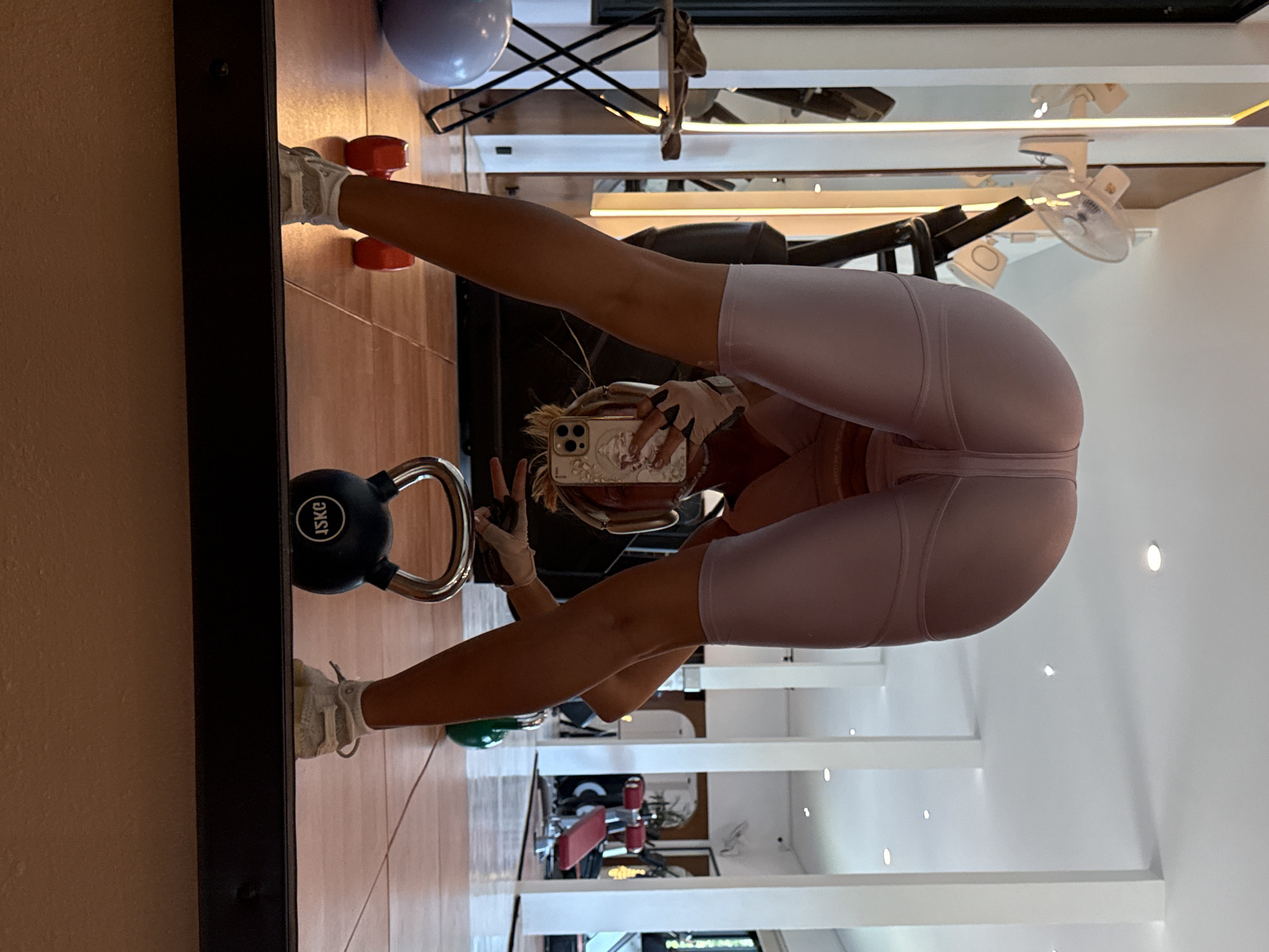 A full-length mirror captures a person in an inverted U-shape pose, possibly stretching or doing yoga, in what appears to be a gym or fitness studio in ตำบลเกาะพะงัน, Thailand. The person is wearing light-colored athletic wear, including shorts and a sports bra, and white sneakers with white workout gloves. They are holding a smartphone in their right hand, suggesting they are taking a mirror selfie.
To the left of the person's feet, a black kettlebell marked "15KG" is visible. Near the person's head, two red dumbbells are on the floor. In the background, reflected in the mirror, various gym equipment can be seen, including what looks like part of a weight bench with red upholstery and other exercise machines. The floor is tiled in a light brown color.
Above the reflections of the gym equipment, ceiling lights and a ceiling fan are visible, indicating an indoor setting. The walls appear to be a light color. The overall impression is that of an indoor fitness environment, likely during daytime given the ambient lighting, although no windows are directly visible. No emotions are discernible from the image.