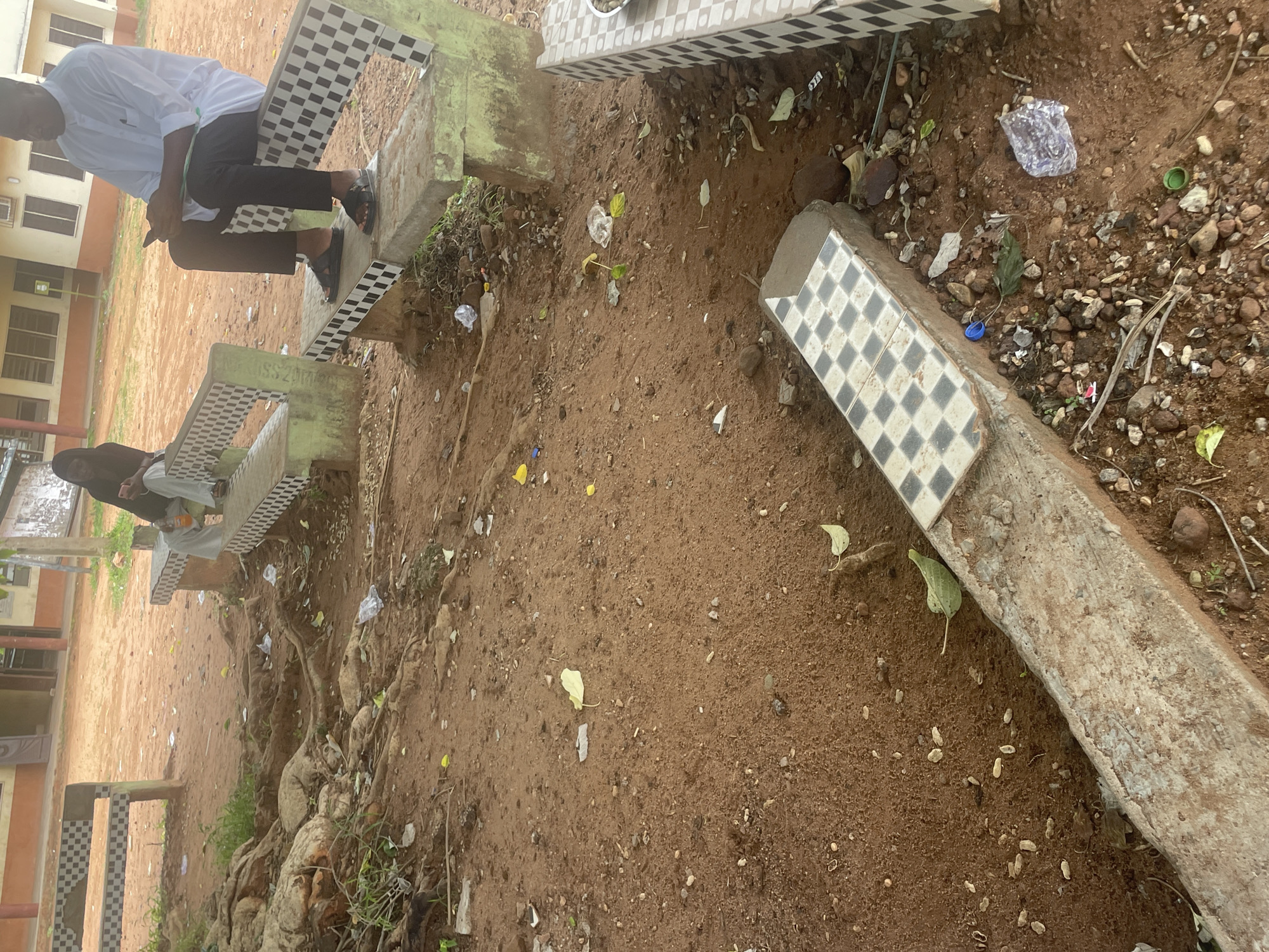 This image depicts an outdoor scene with two individuals seated on a concrete bench decorated with a checkered pattern of black and white tiles. The bench appears weathered, with patches of green paint and exposed concrete. The ground around the bench is composed of red soil mixed with gravel and debris, including fallen leaves and pieces of plastic waste. Large tree roots are visible, suggesting the presence of mature trees nearby.
In the background, a building with multiple windows and orange-colored walls is partially visible. The overall setting appears to be an outdoor seating area, possibly within a school or public space in Lokoja, Nigeria, given the provided location context.
One person, wearing a white collared shirt and dark trousers, is seated on the bench, looking down at their hands, which hold a dark object, possibly a phone. The other person, wearing a dark headscarf and a light-colored garment, is seated on a separate section of the bench, slightly out of focus, with their body turned away from the camera.
The ground is littered with various small items, including plastic wrappers, bottle caps, and scattered pebbles. Fallen leaves are also prominent on the soil. The presence of the debris suggests a less-than-pristine environment, possibly indicating neglect or high foot traffic.
There is no clear indication of the time of day or weather conditions, but the lighting appears natural. No significant emotions are discernible from the subjects' postures. No visible text is present in the image.