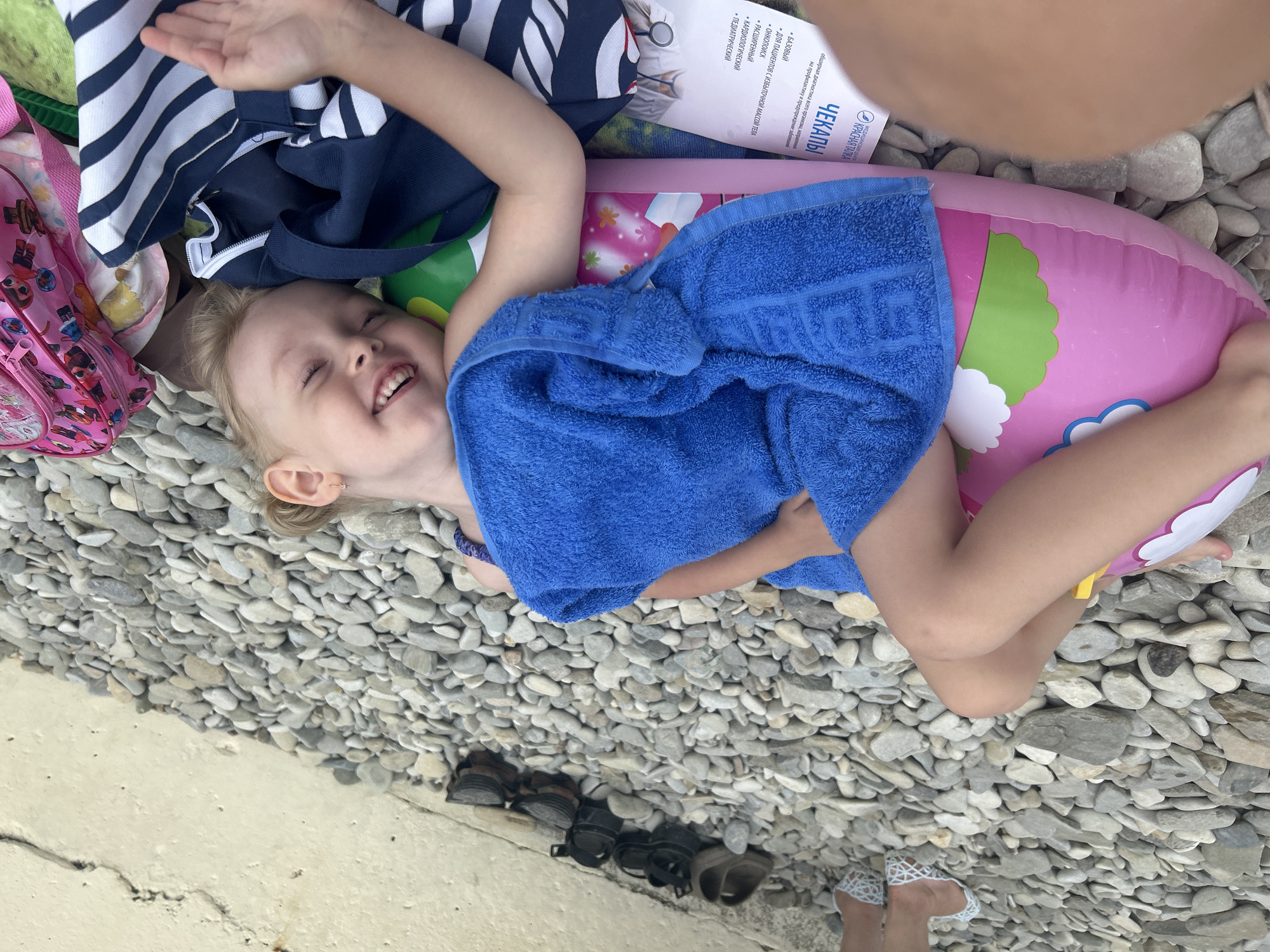 A young blonde child with blue eyes and a wide smile is lying on their back on a pebble beach. The child is wrapped in a blue towel, partially covering a pink inflatable ring. Next to the child is a striped navy and white shirt, and a pink backpack with cartoon characters on it. In the background, there are several pairs of shoes and a concrete wall. The scene is likely the Gelendzhik embankment, a popular tourist destination known for its pebble beaches.