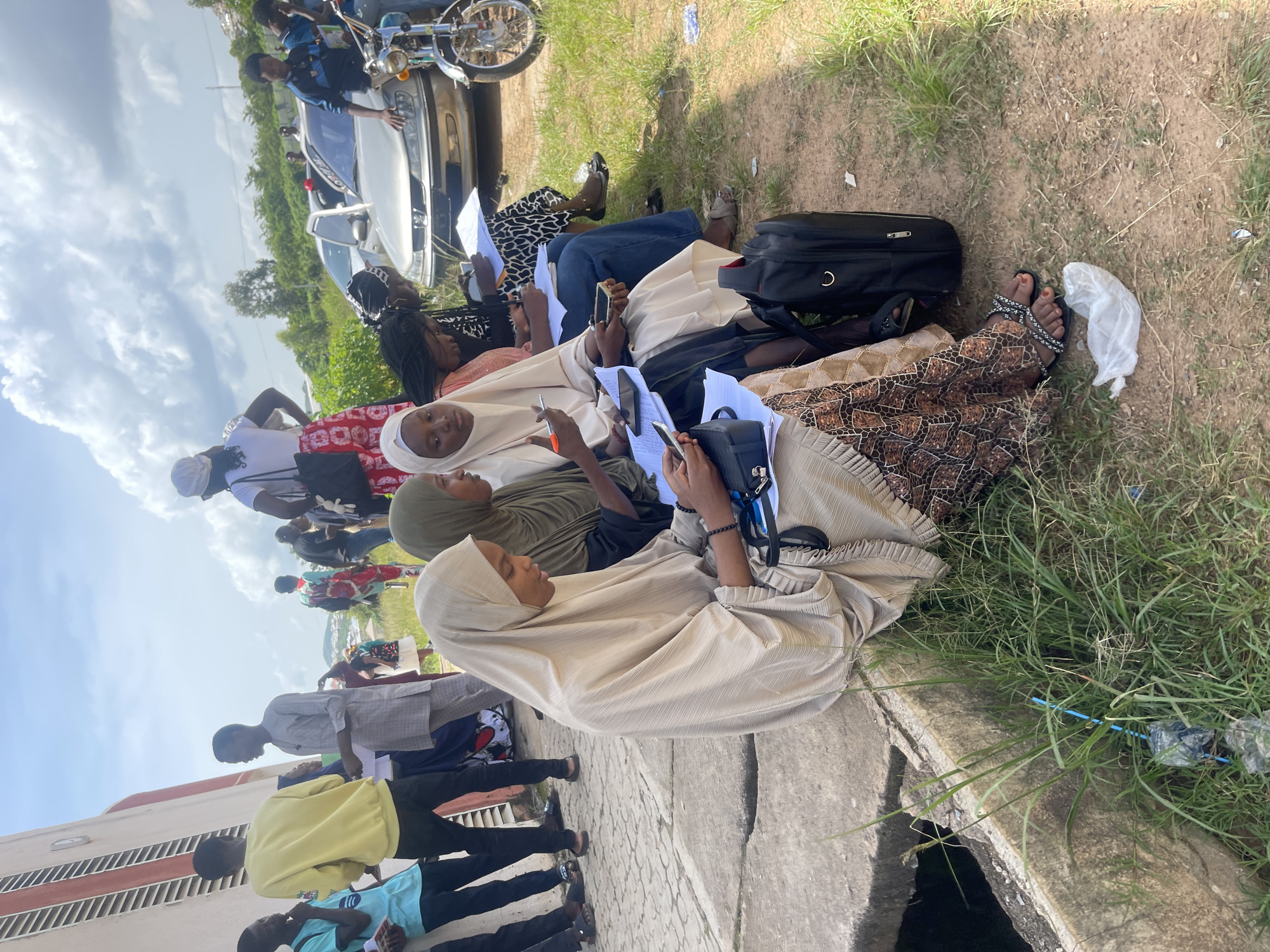 This image depicts a group of young people gathered outdoors on a sunny day, possibly in Jos, Nigeria. In the foreground, several individuals, predominantly young women wearing hijabs and modest clothing, are seated on a concrete ledge. They appear to be engaged in study or examination, with papers and cell phones spread out before them. One young woman in a beige hijab is holding a pen and looking down at her phone, while another to her right is holding a phone and writing on a paper. A black bag rests near them.
Behind this group, several other individuals are standing or sitting. One young man in a yellow sweatshirt is looking towards the left of the frame, interacting with another person. Several other figures are visible in the background, including a person wearing a red and white patterned skirt and white top, and a person wearing a blue and black striped shirt.
The environment suggests an outdoor public space. A silver car is parked behind the seated individuals, and a motorcycle is visible to the right. The ground is a mix of dirt, grass, and a concrete pathway or ledge. In the far background, there are trees and a slightly hazy skyline, with a suggestion of buildings. Some debris, including plastic bags and wrappers, is present on the ground, particularly near the grassy area in the foreground. The sky is bright blue with scattered white clouds. The overall impression is of a casual gathering, potentially related to an educational event or a break in an outdoor activity.