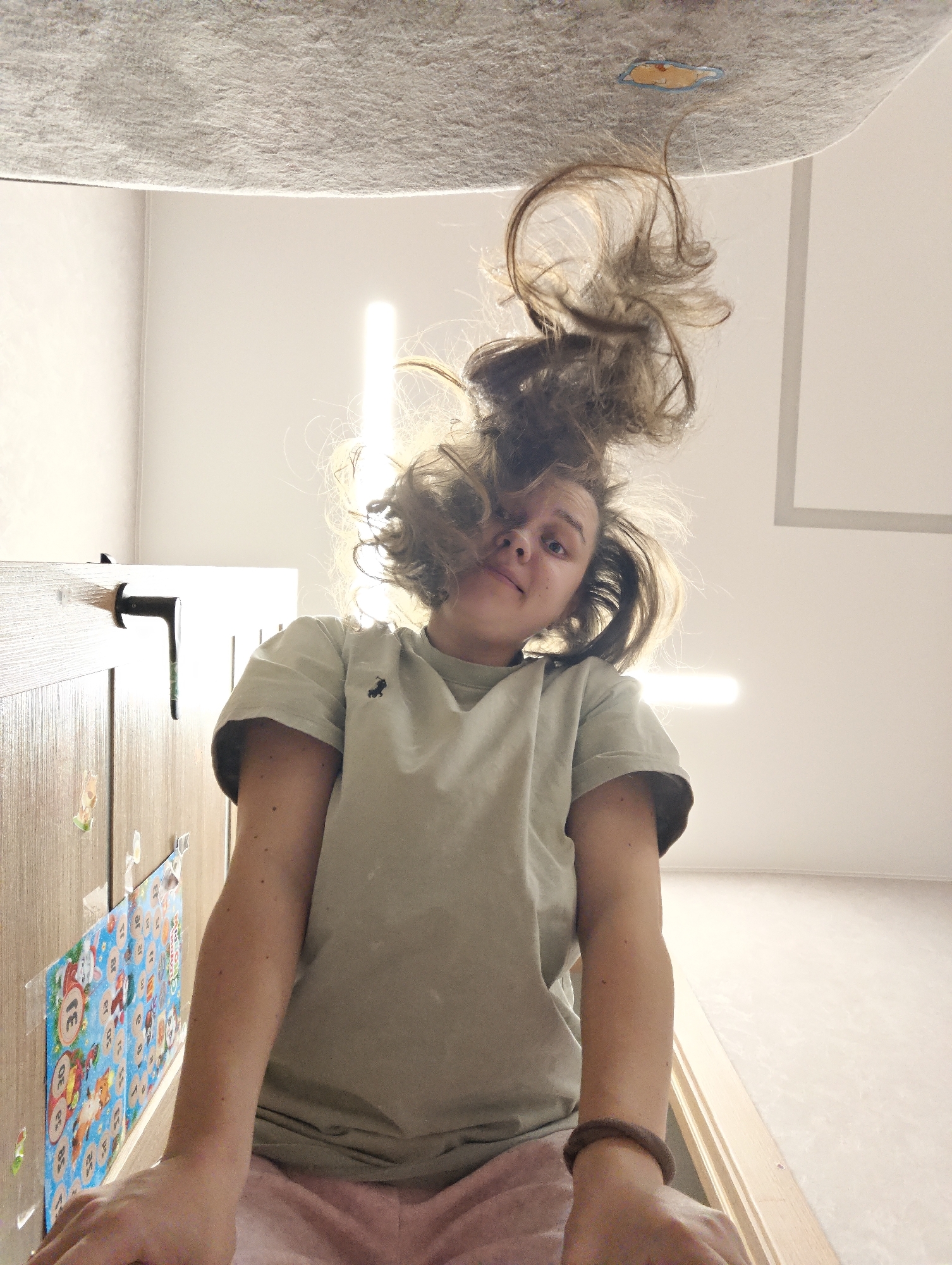 The image shows a woman upside down. Her hair is splayed against a gray ceiling. She is wearing a light green t-shirt and pink shorts. She has a brown hair tie around her wrist. The walls are white and there are fluorescent lights. There is a calendar with cartoon animals attached to a wooden surface in the background. The location is a room somewhere in Chaikovsky, Russia.
