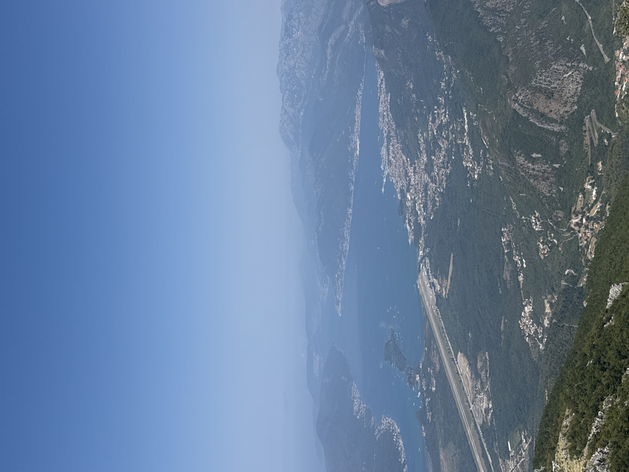 This is an aerial view of a coastal area in Kuk, Montenegro, on a clear, hazy day. The foreground shows steep, rocky, and forested mountainsides with winding roads. In the mid-ground, there is a long, straight airstrip adjacent to a dense green forest. Beyond the airstrip lies a bay with numerous boats scattered across its blue surface. The bay is bordered by a town with buildings clustered along the shoreline, and further in the distance, rolling hills and mountains fade into the haze under a pale blue sky.