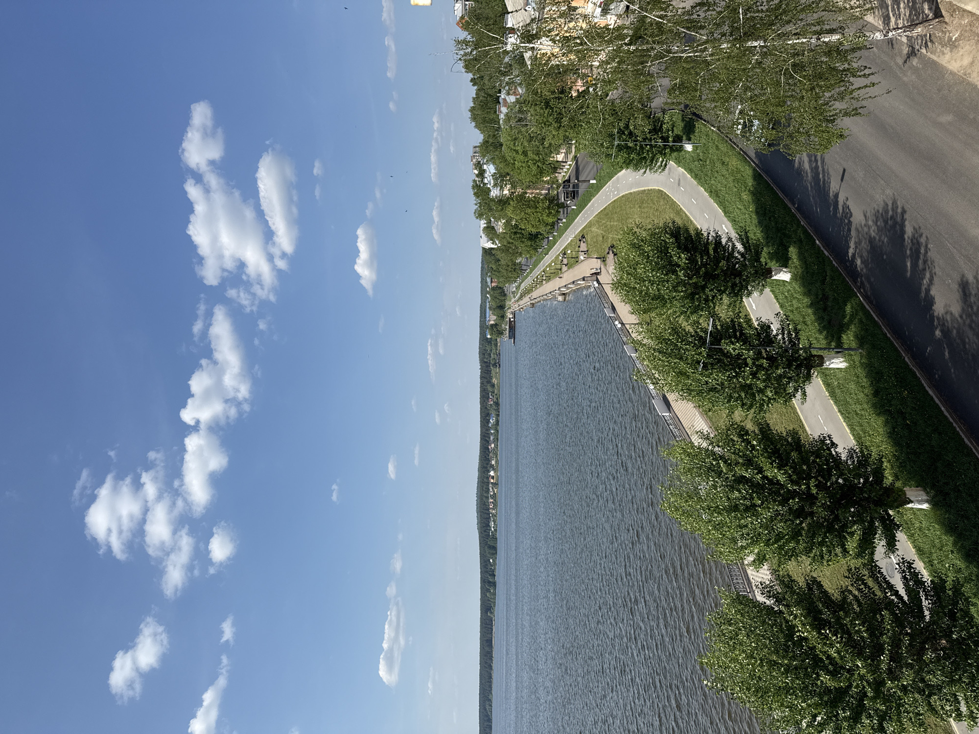 The image depicts a section of the Votkinsk reservoir shoreline in Votkinsk, Russia. A paved embankment runs along the water's edge, lined with trees and benches. A road and a walking/biking path are parallel to the embankment. The reservoir extends into the distance with tree-lined shores on the other side. The sky is blue with scattered white clouds. There are buildings along the edge of the road and behind the trees in the top of the frame. There are no visible people.