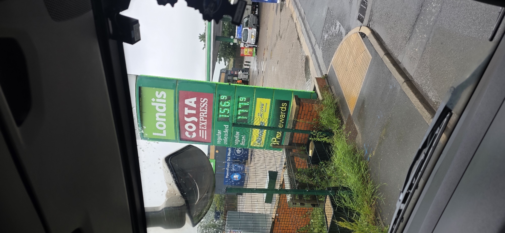 The image depicts a gas station sign with fuel prices displayed. The sign is green and has several panels. The top panel reads "Londis" in white text. Below that, a red panel displays "COSTA EXPRESS". Further down, a green panel shows "regular unleaded" with a price of "156.9" and another panel shows "regular diesel" with a price of "177.9". At the bottom of the sign, a green panel reads "3p me ewards".
In the background, there is a canopy over the gas pumps. There are several vehicles, including cars and vans, at the station. There are also some individuals, possibly gas station employees, wearing orange vests. To the left of the sign, there is a car wash facility.
The ground is wet, indicating it may have rained recently. The scene appears to be set in an outdoor environment, likely a convenience store or gas station. The image is taken from inside a vehicle, with a windshield wiper visible in the foreground. The location is identified as Tameside, United Kingdom.