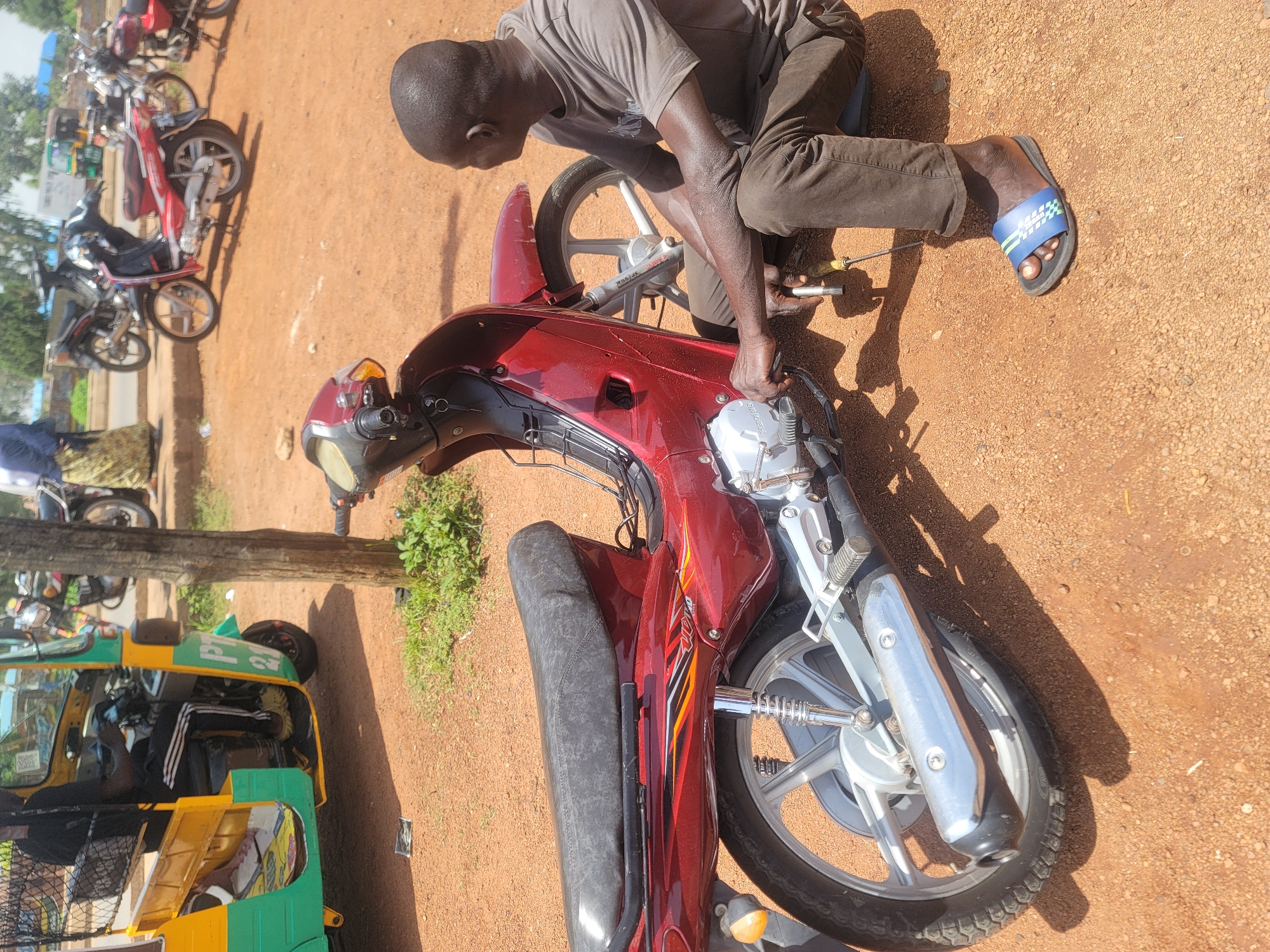 In Barakin Lerawi, Nigeria, a man is seen crouched down next to a maroon motorcycle, appearing to be performing maintenance. He is wearing a dark grey t-shirt and dark brown trousers. His right hand is on the motorcycle's engine area, and he holds a tool. A screwdriver lies on the ground near him. The motorcycle is parked on a reddish-brown dirt surface. In the background, a green and yellow auto-rickshaw is visible, along with several other motorcycles parked along a road. A tree trunk stands to the left of the motorcycle. The environment suggests an outdoor, potentially informal workshop or roadside setting within the town.