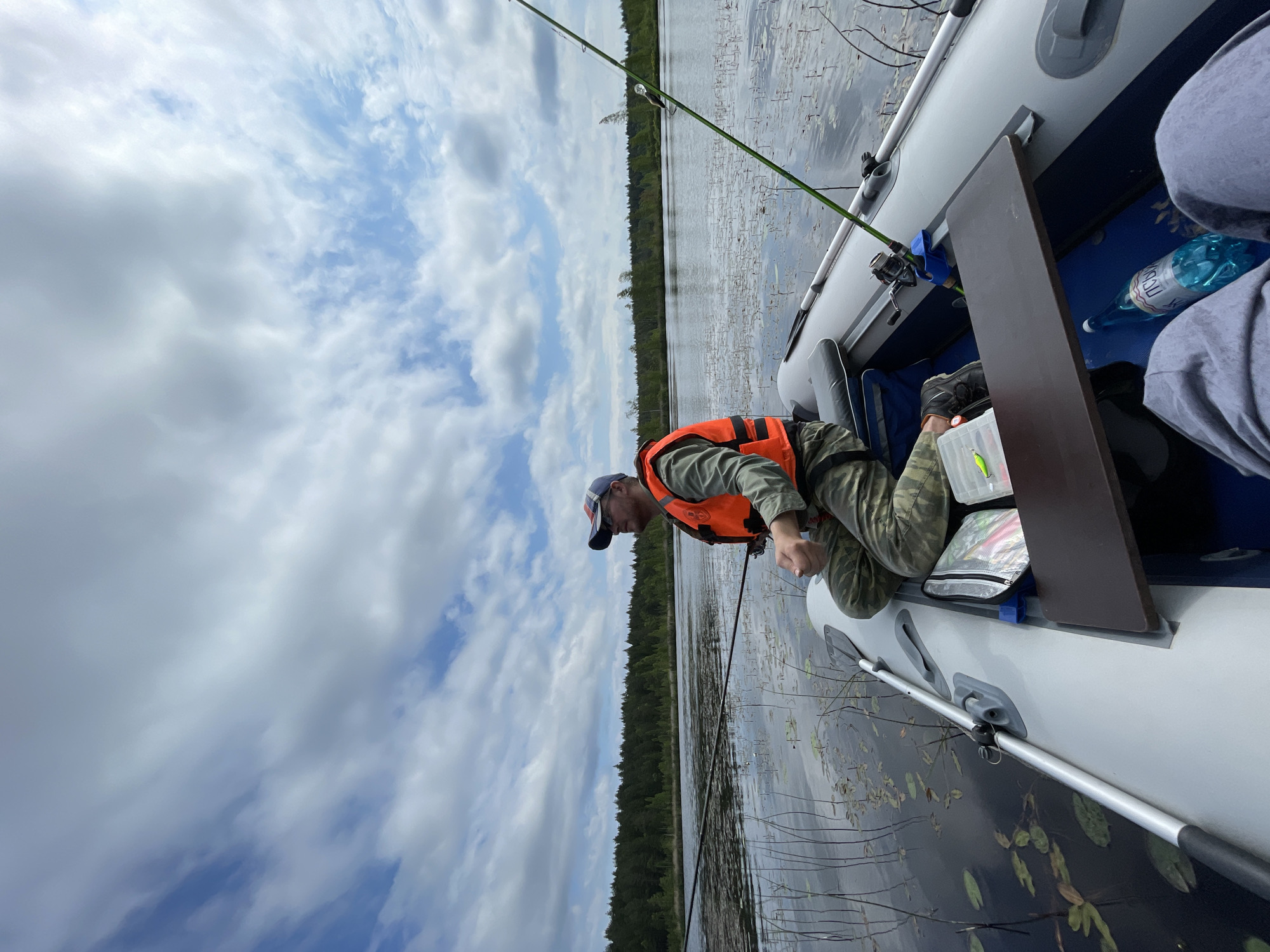The image depicts a man fishing from an inflatable boat on a lake. The man is wearing a camouflage baseball cap and an orange life vest over a green long-sleeved shirt and camouflage pants. He is seated in the boat, holding a fishing rod. The boat contains fishing gear, including a tackle box with lures and a bottle of water. The lake is surrounded by a dense forest. The sky is partly cloudy with white and gray clouds. The location is identified as Roshchino, Russia.