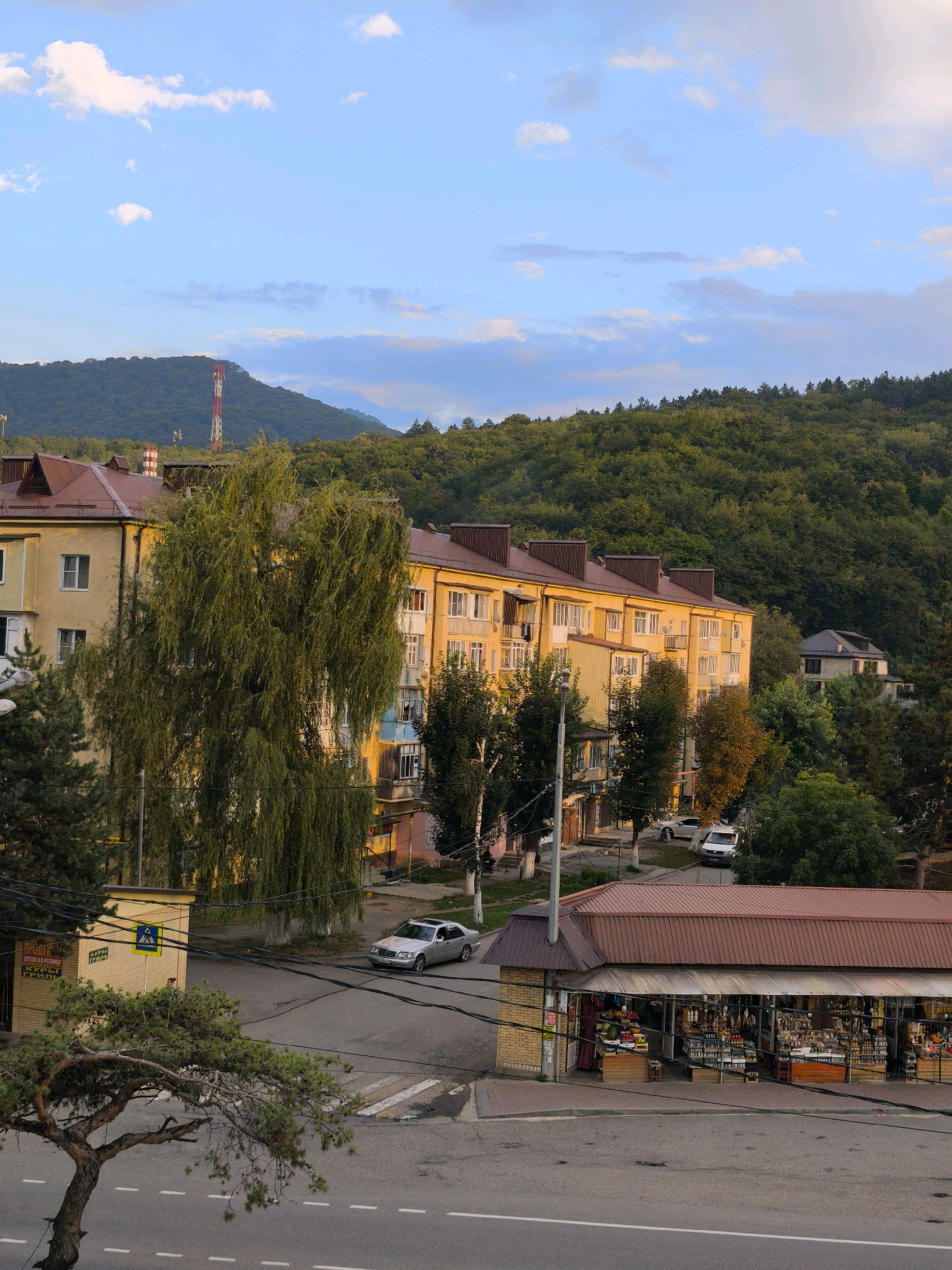 The image captures a street scene in Karachayevsk, Russia, during daylight. In the foreground, a small market stall with a reddish-brown roof is visible, displaying various goods such as bottles, produce, and dried items. Adjacent to the stall, a yellow building with a sign featuring Cyrillic text is situated. A crosswalk is marked on the asphalt road. A silver sedan is parked on the street, and a white van is also present. Further down the road, a multi-story residential building with yellow walls and dark brown roofs stands prominently, with a large weeping willow tree in front of it. Behind the buildings, a dense green forest covers the slopes of a mountain range. A red and white radio tower is visible on one of the mountain peaks, and a light blue sky with scattered clouds completes the scene. The overall impression is a quiet urban environment nestled against a natural landscape.