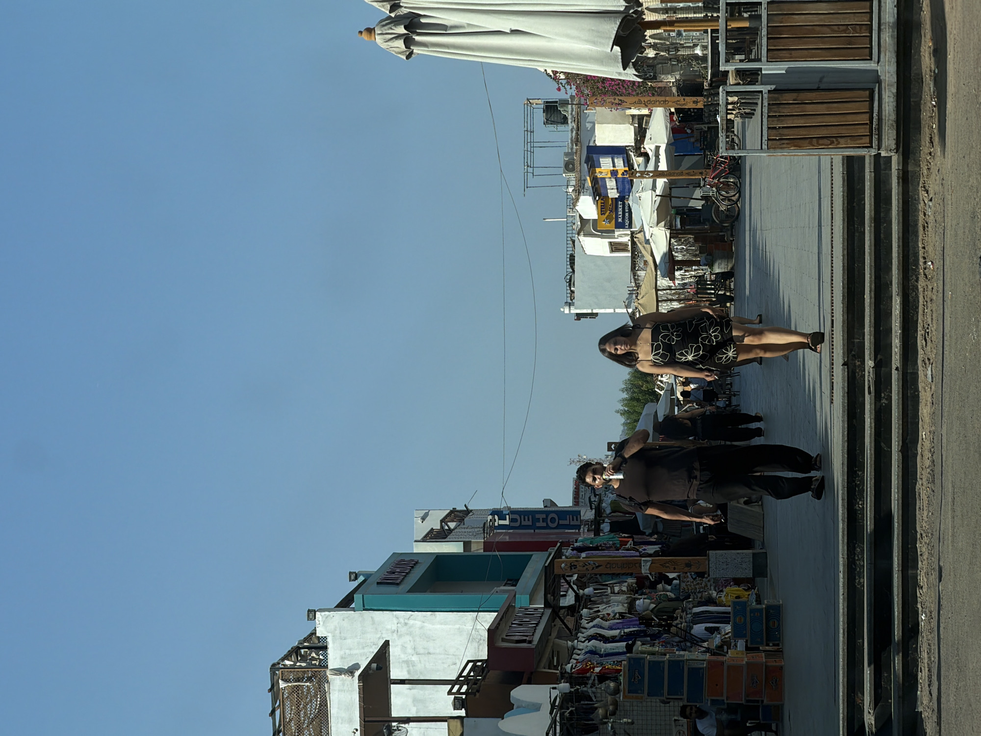 The image depicts a street scene in Dahab, Egypt. A man in dark clothing is walking in the foreground, holding a can to his mouth. To his right, a woman in a black and white floral dress is walking. Shops with awnings and clothing displays are visible on the left side of the street, with signage indicating businesses. Umbrellas and tables are set up outside some establishments on the right. In the background, buildings with flat roofs and air conditioning units are visible against a clear blue sky. Several bicycles are parked near a shop with a yellow and blue sign. The street appears to be paved, and there are steps leading down from the sidewalk in the foreground.