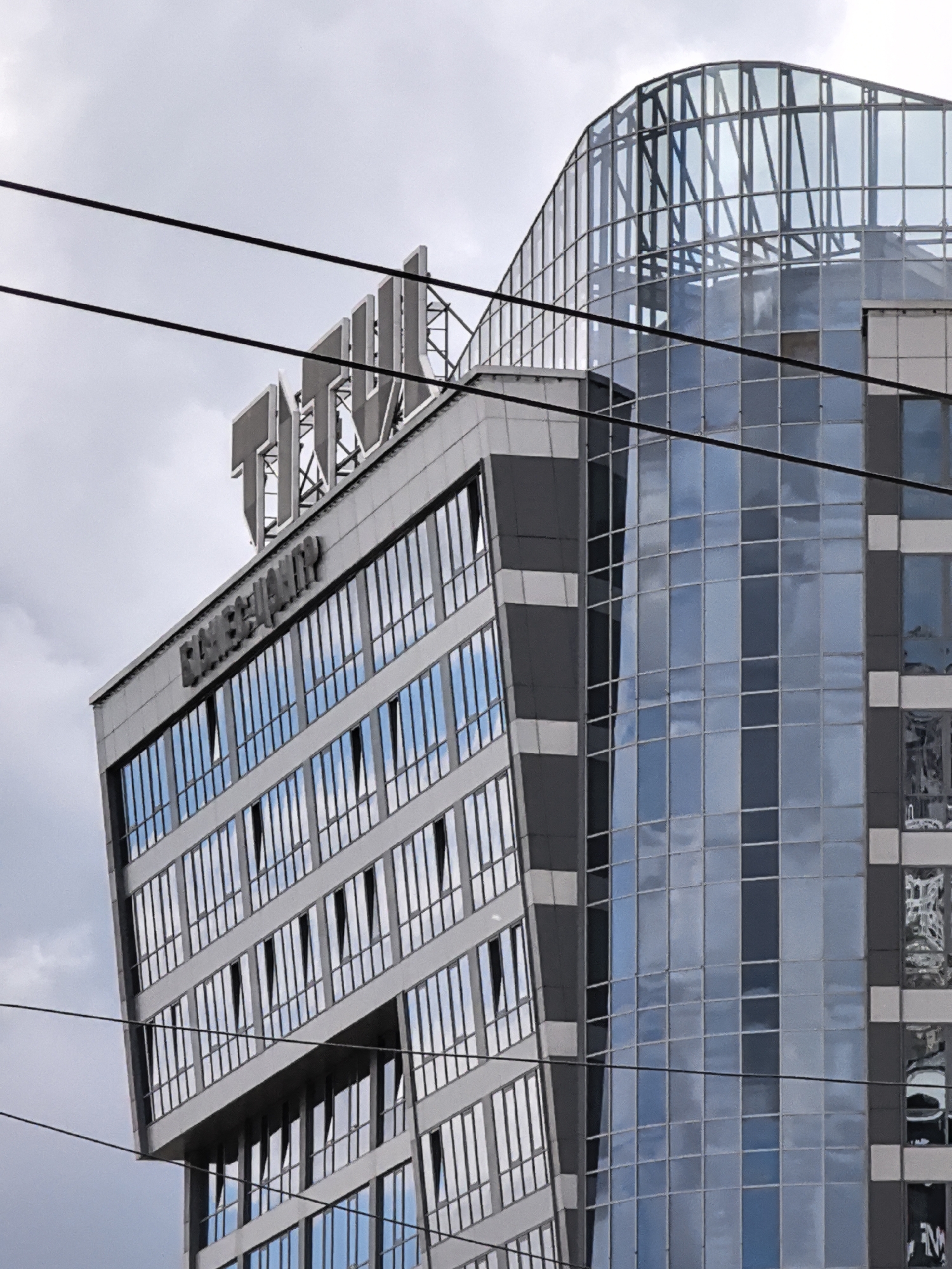 A low-angle, full shot shows the upper floors of a modern, glass-fronted office building on an overcast day. The building is tall and angular, with sections of grey and dark grey siding interspersed with large glass windows. A curved section of glass rises from the top of the building on the right side. The building has a large, three-dimensional sign on its roof, with letters that appear to be made of metal. Below this, a smaller sign with dark letters reads "ВАСІЛЬЄВ". Several black electrical wires are strung across the foreground, obscuring parts of the building. The sky is cloudy and grey.