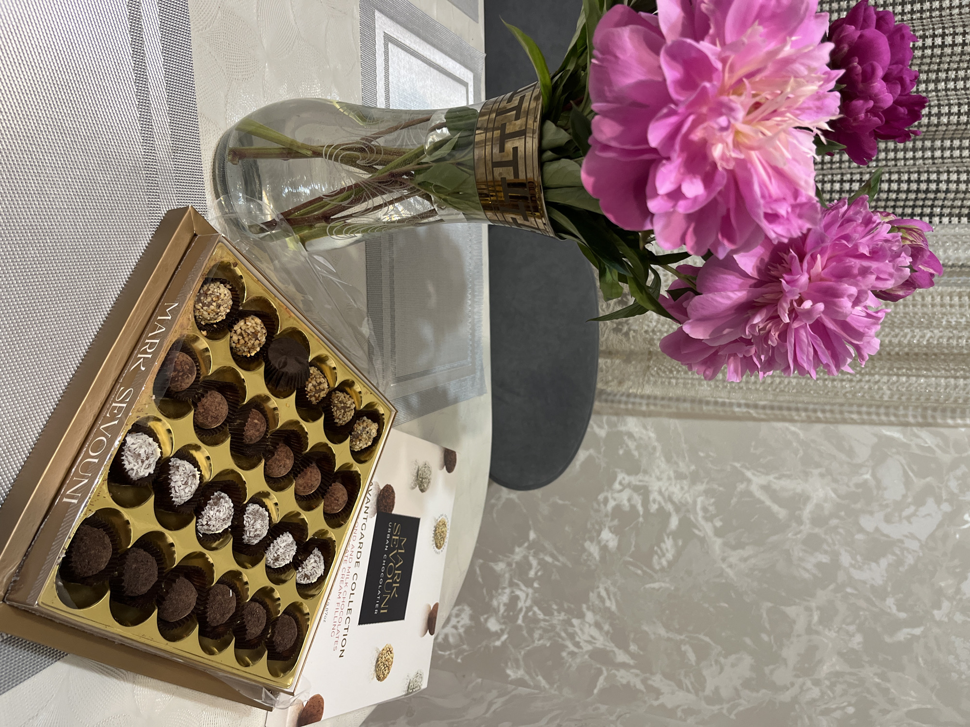 The image depicts a box of Mark Sevouni Avantgarde Collection chocolates alongside a vase of pink and purple peonies, placed on what appears to be a table with a patterned surface. The chocolate box is positioned at the left of the image and has an open lid revealing various types of chocolate truffles. The vase, containing water and supporting the peonies, sits to the right of the chocolate box, with the flowers occupying the upper right portion of the frame. The table's surface has a white and gray marble-like pattern. The location, Kaspiysk, Russia, is not directly discernible from the visual elements of the image alone.