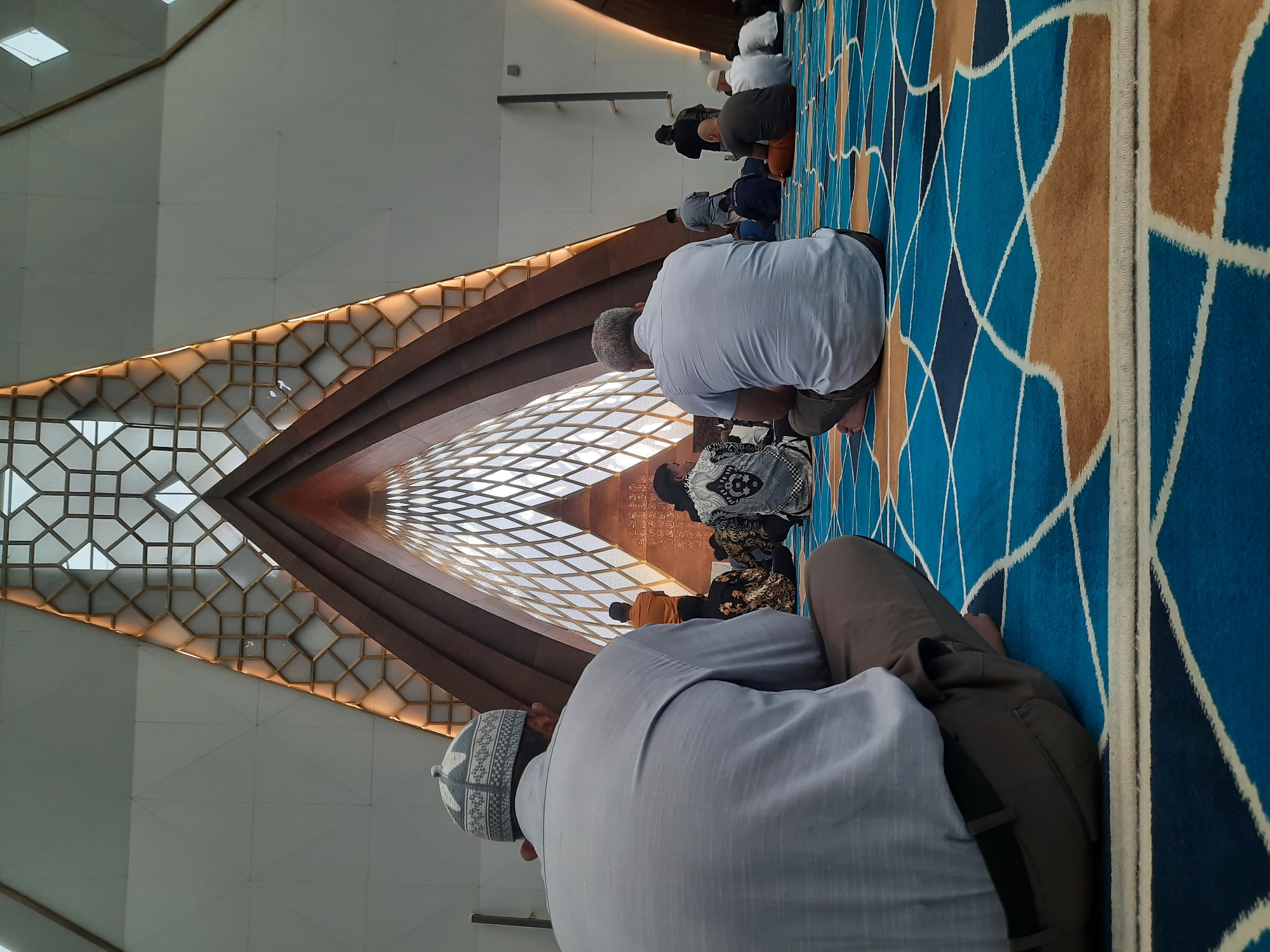 The image depicts the interior of a mosque in Bandung City, Indonesia, during a prayer service. The perspective is from behind a worshipper in the foreground, seated on a blue and gold patterned carpet. The mosque features a striking architectural design with a large, vaulted ceiling that tapers upwards, adorned with intricate geometric patterns and wooden accents. Natural light filters through a large, diamond-shaped window at the apex of the ceiling, illuminating the space. Several other individuals are seen in prayer or seated on the carpet throughout the mosque. The overall atmosphere is one of quiet reverence and communal worship.