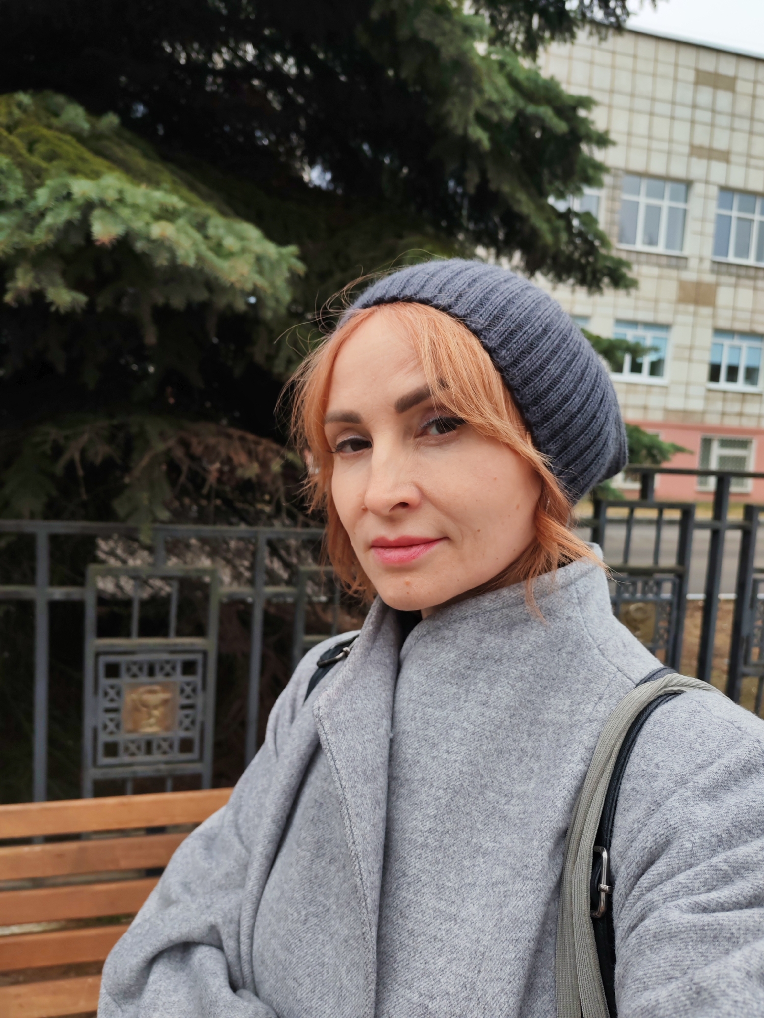 A woman is captured in a close-up outdoor portrait in Chaikovsky, Russia, on what appears to be an overcast day. She is looking directly at the camera with a neutral to slightly content expression. She has light reddish-blonde hair, styled with bangs, and is wearing a dark grey knit beanie and a light grey wool-blend coat. A dark bag strap is visible over her right shoulder.
The background features elements of an urban or park setting. To the woman's left, part of a wooden slatted bench is visible. Behind her, a dark grey metal fence with decorative, possibly gilded, square panels runs horizontally across the frame. Dense green coniferous trees dominate the upper left and central background, suggesting a natural or planted area. On the upper right, a light-colored, multi-story building with numerous rectangular windows and a grid-patterned facade can be seen, possibly an institutional or residential structure. The soft, diffused lighting indicates an overcast sky, complementing the cool tones of the woman's attire and the wintery/autumnal feel conveyed by the heavy coat and hat. There is no visible text in the image.