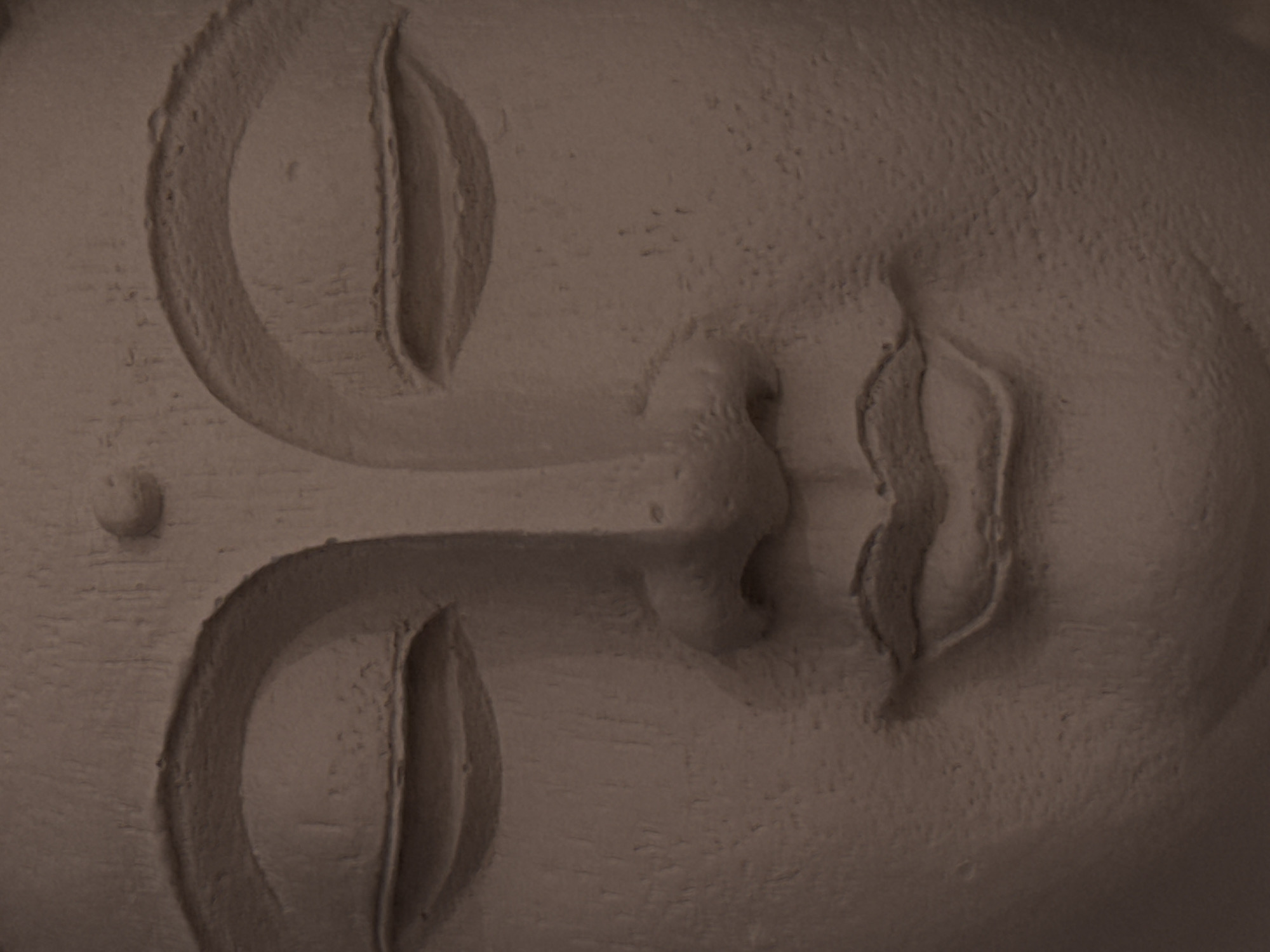 The image is a close-up, low-angle shot of a sculpture's face. The sculpture appears to be made of a textured, matte material, possibly clay or stone, with a subtle roughness that catches the light. The dominant color is a muted, earthy brown. The focus is on the facial features, with particular detail given to the closed eyes, defined by sculpted eyelids and prominent brow ridges. The nose is broad and rounded, and the lips are full and gently curved, suggesting a serene or meditative expression. The texture of the material creates shadows and highlights that emphasize the contours of the face. The background is indistinct, blending with the sculpture itself, and suggesting it's a studio or a display setting rather than an outdoor environment. There is no indication of location in Moscow, Russia, or any discernible environment beyond the sculpture itself.
