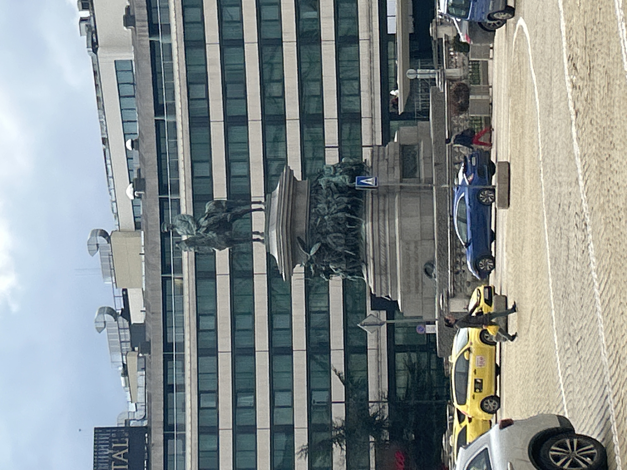 The image shows a street scene in Sofia, Bulgaria. A large building with a grid of windows forms the backdrop. In front of the building, there's a monument featuring a horse-mounted statue. To the right, there's a street with several parked vehicles, including a yellow taxi. A person appears to be leaning against the taxi. The environment includes a sidewalk, pavement, and street-level features like streetlights and signage.