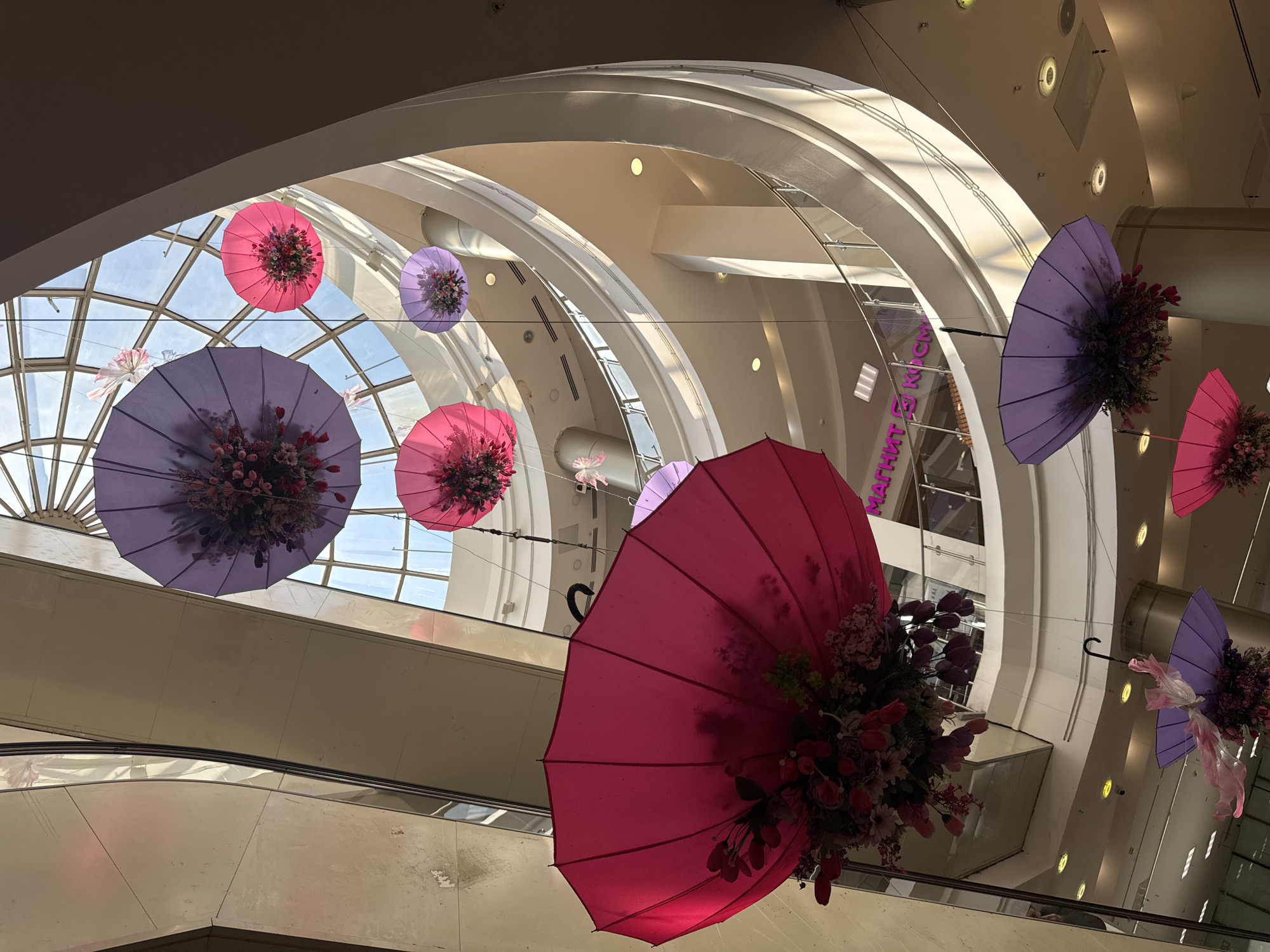 The image shows an interior view of a shopping mall in Kazan, Russia. The ceiling features a combination of arches and glass panels, allowing natural light to flood the space. Decorative elements include multiple open umbrellas suspended from the ceiling at varying heights, each adorned with floral arrangements. The umbrellas are in pink and purple shades. An escalator is visible in the background.