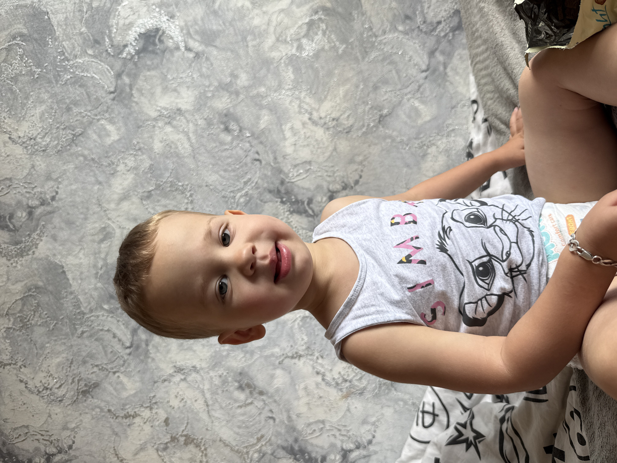 A young Caucasian child, appearing to be between two and three years old, is seated on a bed against a textured grey wall in a residential interior, likely in Rusanovo, Russia. The child has light brown hair and blue eyes, and is wearing a light grey tank top with a Simba graphic from The Lion King and a diaper. They are looking directly at the camera with a slight smile. The bed is covered with patterned black and white bedding and a grey blanket. To the right of the child, a partially visible package of dark chocolate or cookies rests on the blanket. The overall lighting suggests an indoor setting during daylight.