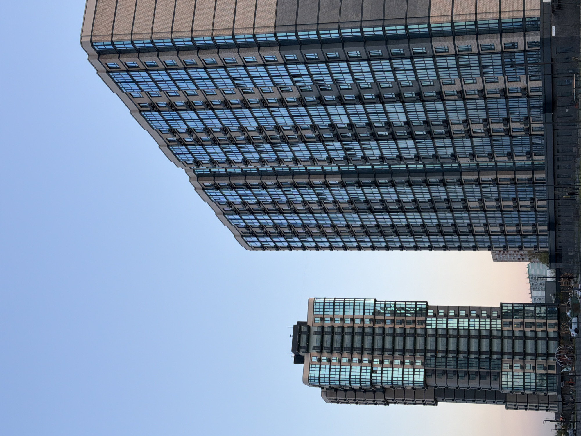 Two modern, multi-story buildings dominate the urban landscape against a clear sky. The larger building, positioned on the left, features a light brown or beige brick-like facade with numerous rectangular windows set in dark frames. Many of these windows reflect the blue sky, and some appear to be open. The building's design includes horizontal banding and a large, expansive wall of windows.
To the right, a second, slightly smaller building stands. It shares a similar architectural style with a mix of light-colored panels and dark-framed windows, also reflecting the sky. This building exhibits a more stepped or terraced structure, giving it a distinctive silhouette.
The sky, transitioning from a light blue at the top to a faint, soft pinkish-orange near the horizon, suggests either an early morning or late afternoon setting. No clouds are visible, indicating clear weather. In the background, between the two main structures, other less distinct buildings can be seen under the lighter horizon. The overall scene depicts a dense urban environment in Yuzhny, Russia, showcasing contemporary architectural styles. There is no visible activity or legible text.