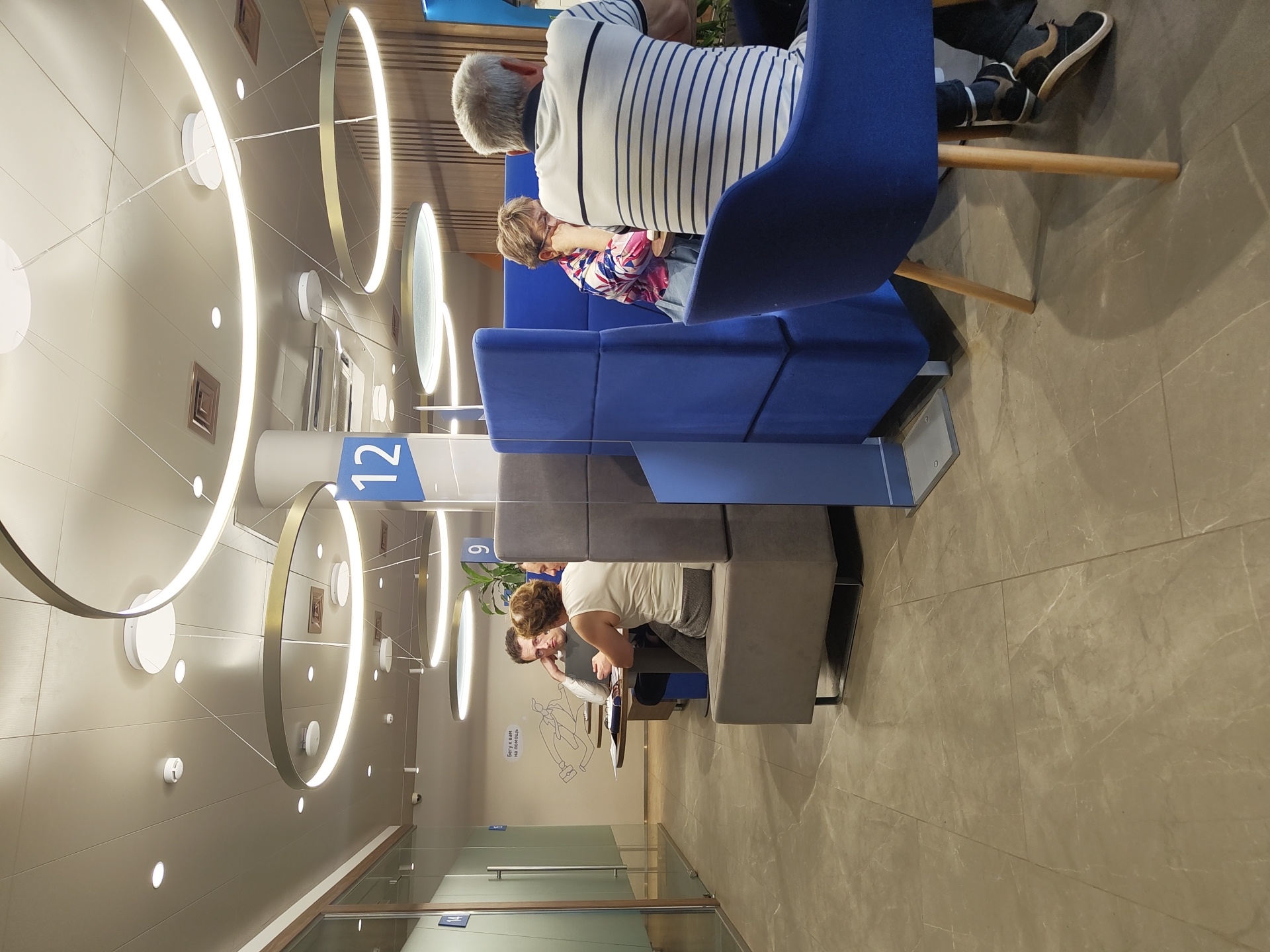 The image depicts the interior of what appears to be a modern bank or office waiting area in Ramenskoye, Russia. The scene is brightly lit by contemporary circular pendant lights and recessed ceiling lights. In the foreground, a man with gray hair, wearing a striped t-shirt and dark pants, is seated in a blue armchair, facing away from the camera. To his left, within a blue partitioned seating area, a woman with short, light brown hair, wearing a colorful patterned shirt, leans on her hand, looking down. Further back, on the left side of the image, a man and a woman are seated at a table, engaged in conversation. The man, wearing a white shirt, is looking at the woman, who is wearing a white top and has her back to the camera. The environment features beige tiled flooring, gray and blue upholstered seating booths, and glass partitions. A large blue sign with the number "12" is prominently displayed on a white pillar. The walls are decorated with modern artwork and a potted plant is visible. The overall impression is one of a clean, organized, and somewhat corporate space designed for client waiting and interaction.