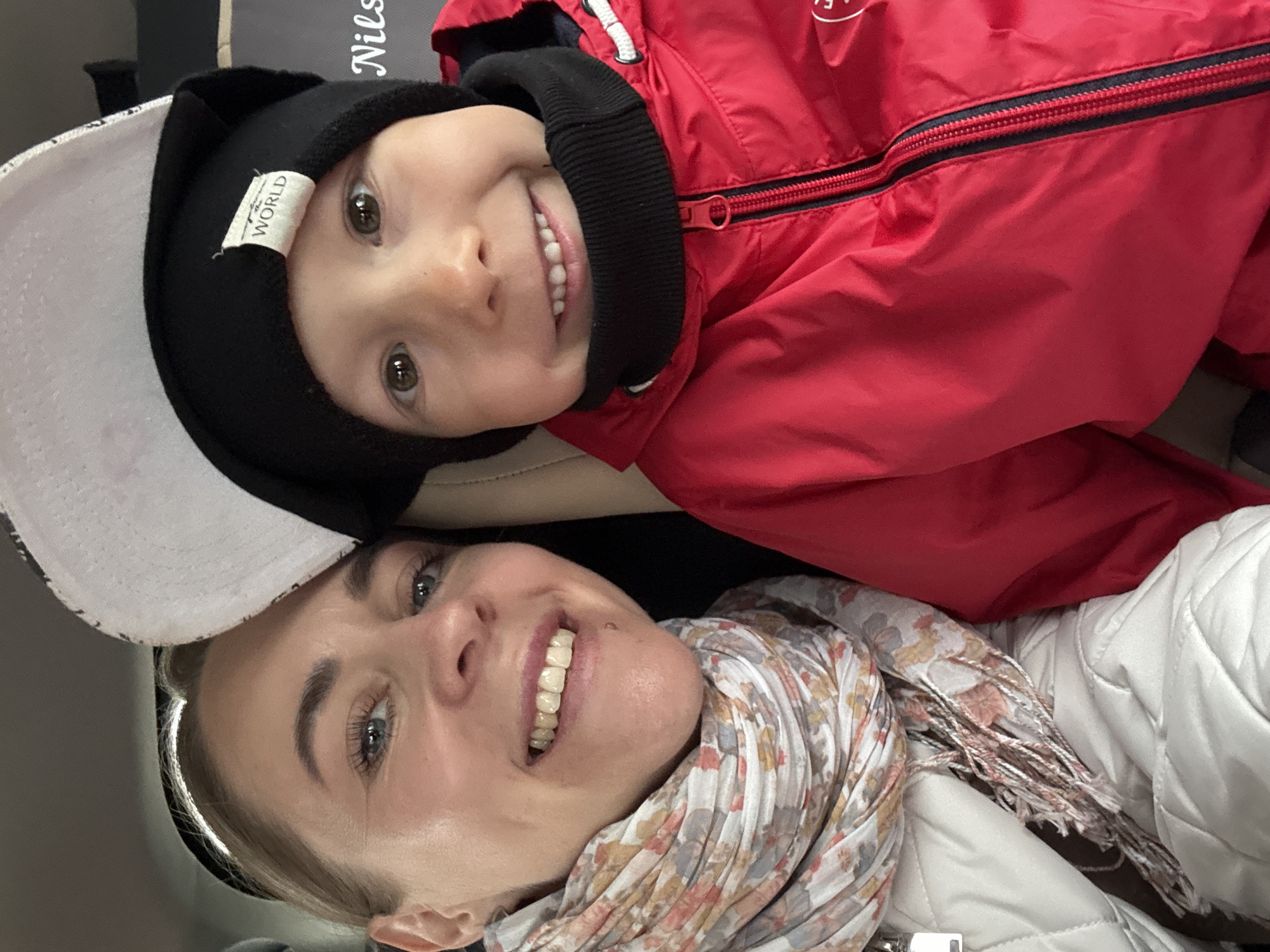 The image shows a selfie of a woman and a young child, likely indoors. The woman is wearing a white quilted jacket and a floral scarf, and a hat. The child is wearing a red jacket and a black beanie. Both are smiling at the camera. The location is unknown, but the user requested that Chaikovsky, Russia, be included. There are no indicators within the image that corroborate this.