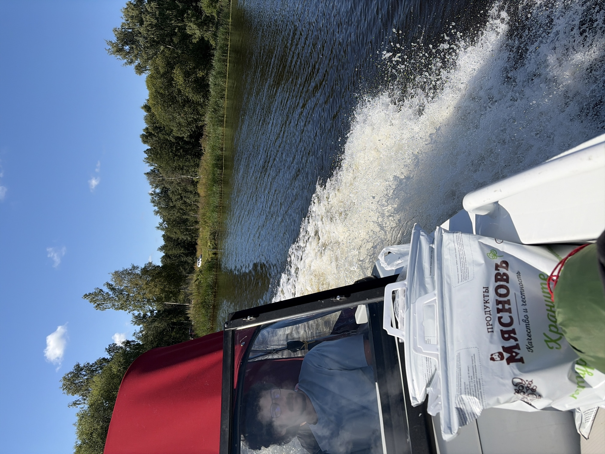 A person with dark, curly hair and glasses is driving a boat on a dark blue body of water. They are wearing a light blue t-shirt and are positioned behind a clear windshield. The boat has a red canopy extending from the top. In the foreground of the boat, there are two large white bags with handles, one stacked on top of the other. The bags have text printed on them, including the words "ПРОДУКТЫ" and "МЯСНОВЪ". The wake of the boat is visible trailing behind it, with white foam and splashes. On the right side of the image, a shoreline is visible with a dense line of green trees and tall grass. The sky is clear and blue with a few scattered white clouds. The scene appears to be a sunny day on a body of water, possibly a river or lake, in a natural setting. The specific location in the city of Novomelkovo, Russia, is not discernible from the image itself.